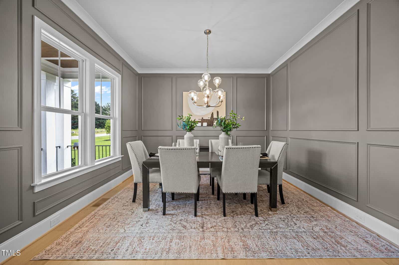 A stylish dining room with gray paneled walls, a rectangular glass table surrounded by six upholstered chairs, a modern chandelier overhead, fresh flowers on the table, a large window, and a patterned rug on hardwood floors.