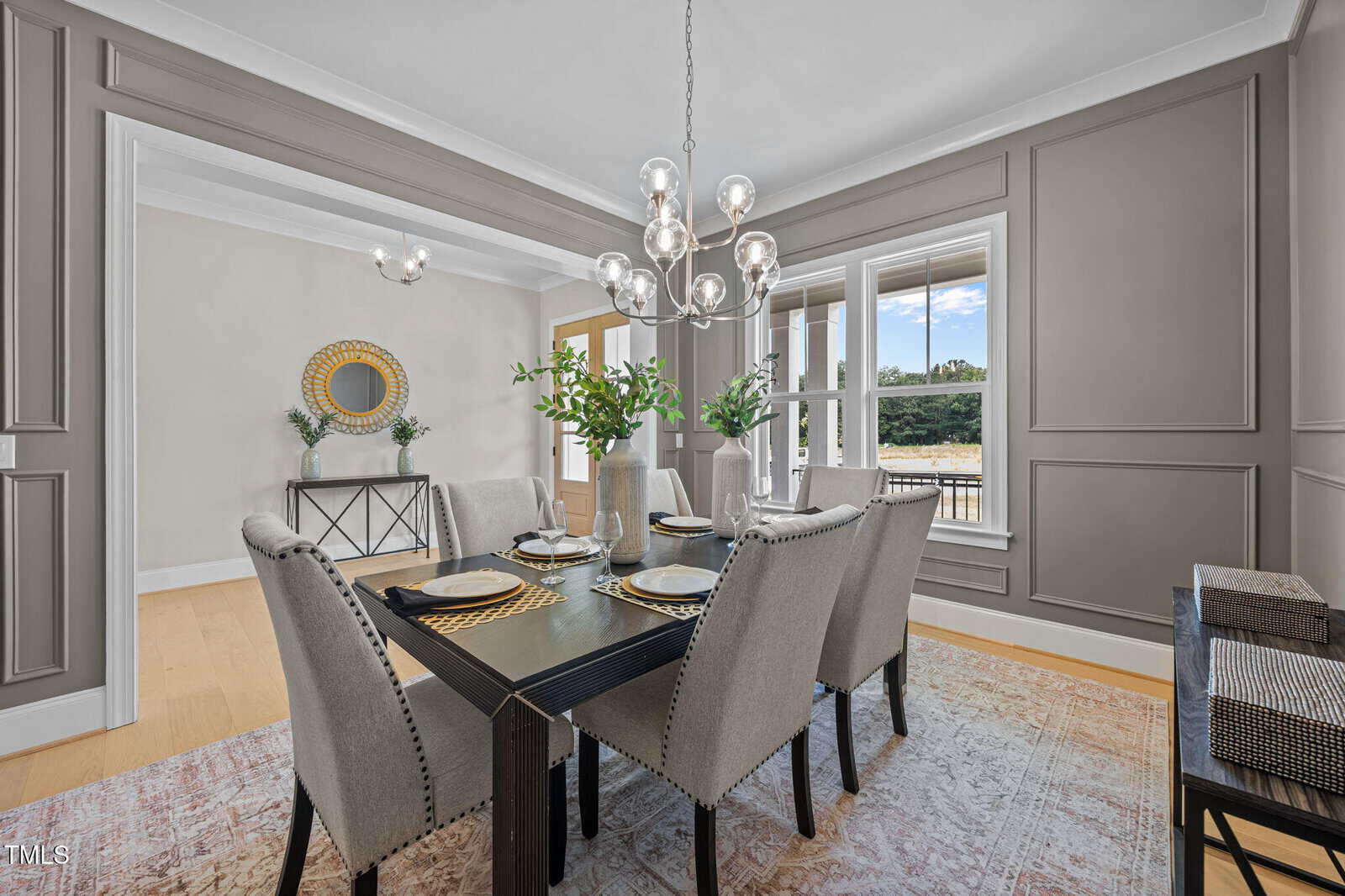 A modern dining room with a dark rectangular table set for six, gray upholstered chairs, two vases with greenery, a chandelier, large windows, gray paneled walls, and a console table with a round mirror.