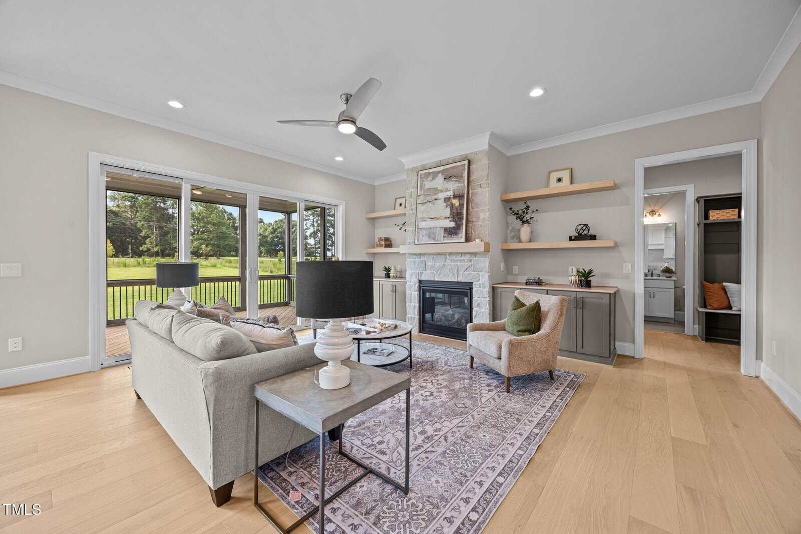 Modern living room with light wood floors, neutral furniture, built-in shelves, a stone fireplace, and large windows overlooking a green backyard. A ceiling fan and recessed lights are also visible.