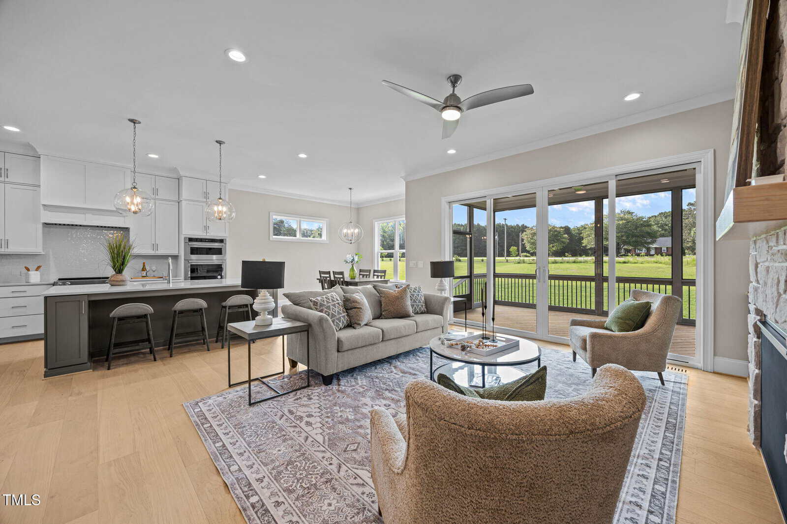 Modern open-concept living room and kitchen with neutral tones, large windows, ceiling fan, and view of a green backyard. Includes a sofa, two chairs, and a kitchen island with pendant lights.