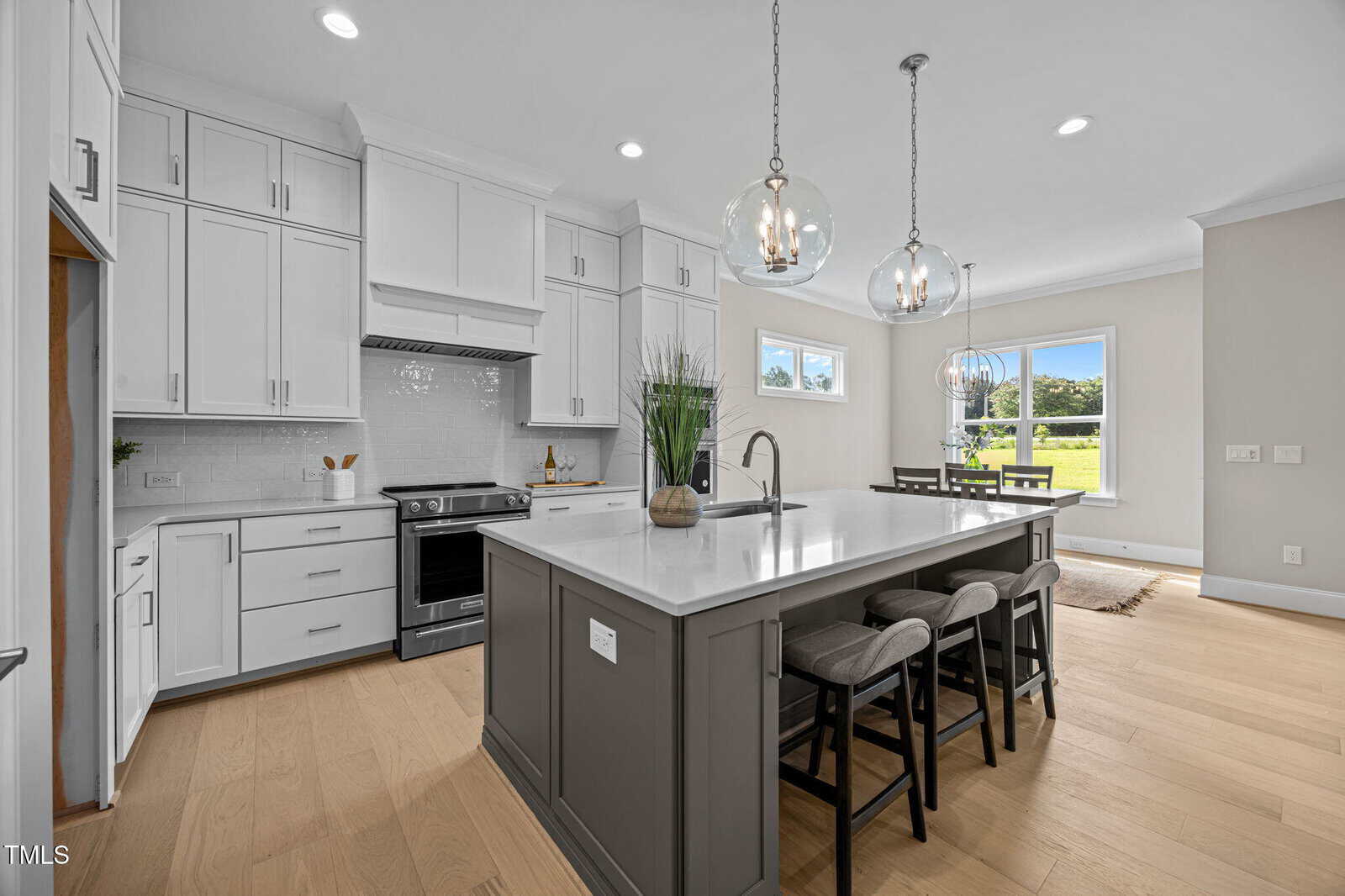 Modern kitchen with white cabinets, stainless steel appliances, a large gray island with sink, three upholstered bar stools, pendant lights, wood floors, and large windows letting in natural light.