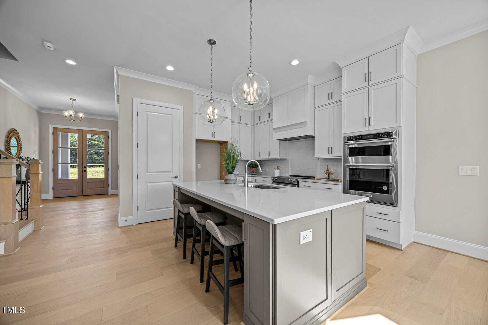Modern kitchen with white cabinets, stainless steel appliances, a large island with barstools, pendant lights, and light wood flooring. A hallway with double front doors is visible in the background.
