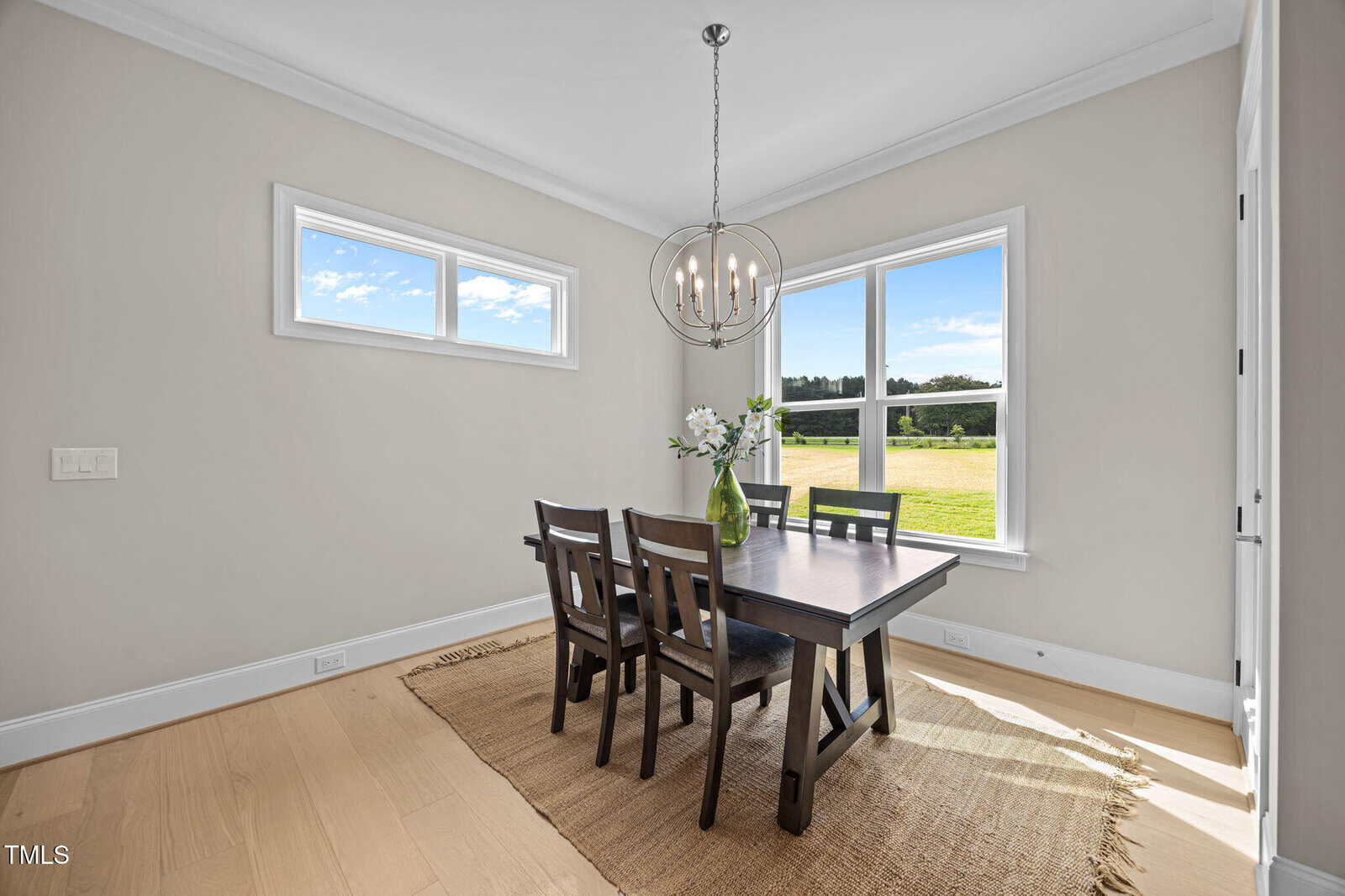 A bright dining room with a dark wooden table, four chairs, a vase with flowers, large windows letting in sunlight, light wood floors, a beige rug, and a modern chandelier. The windows show a grassy outdoor view.