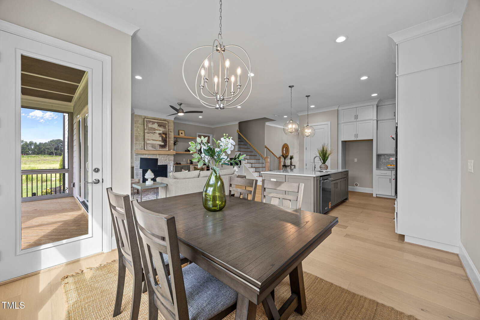 Modern open-concept kitchen and dining area with wooden table, green vase centerpiece, chandeliers, island with stools, and view of a furnished living room. Glass door leads to a screened porch and green outdoor scenery.