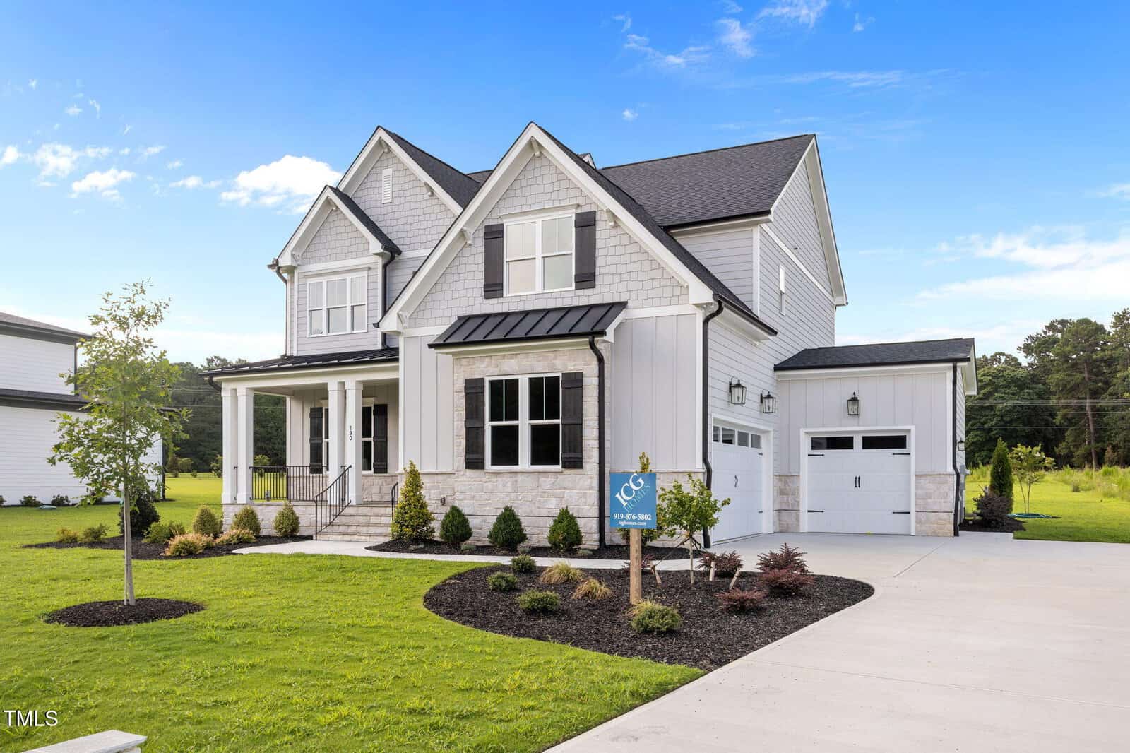 A two-story modern suburban house with white siding, black shutters, and a covered front porch, surrounded by a well-manicured lawn, young trees, and neatly landscaped shrubs under a blue sky.