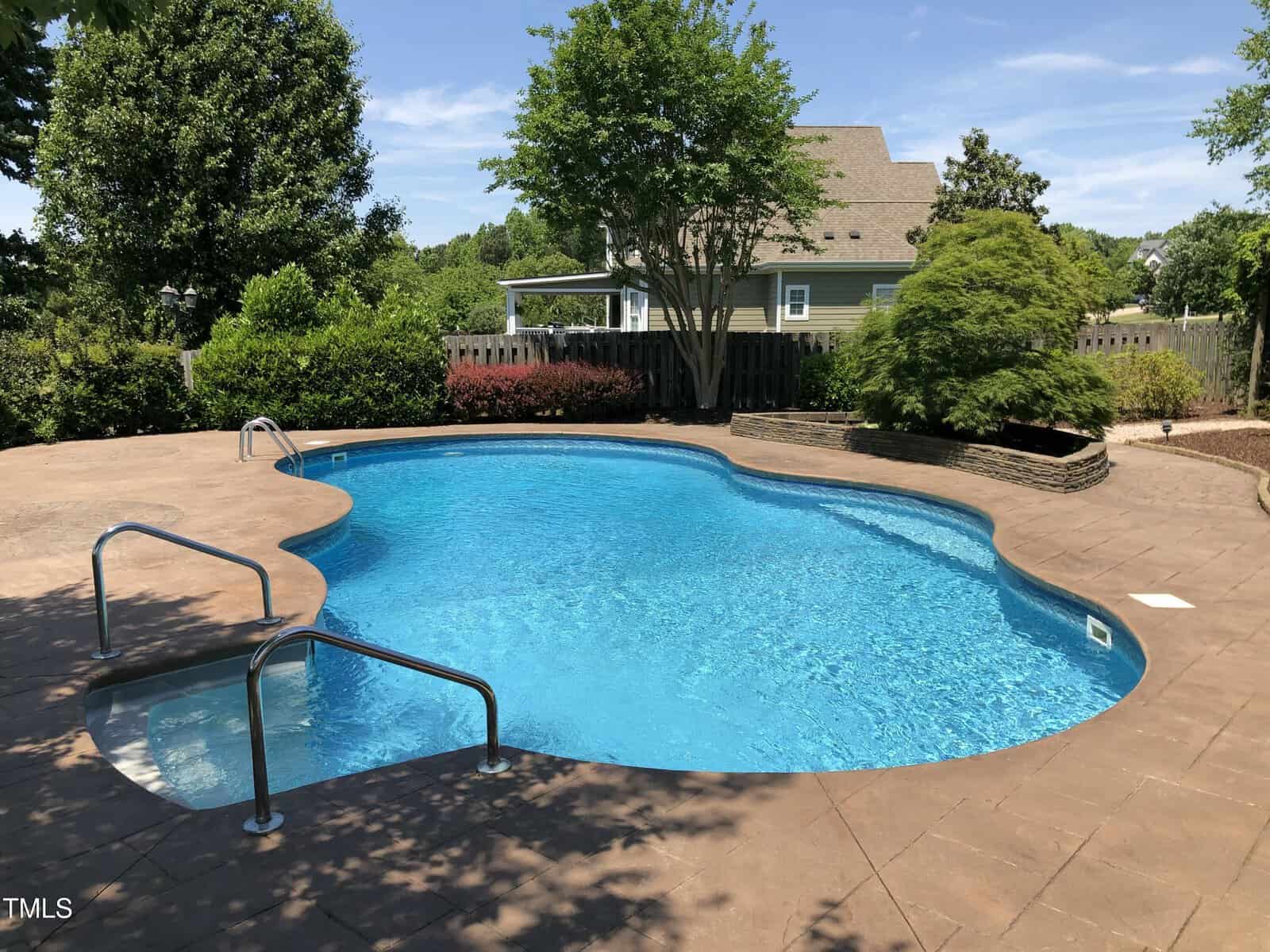 A clean, kidney-shaped backyard pool with metal handrails, surrounded by a brown concrete deck, green trees, bushes, and a house in the background on a sunny day.