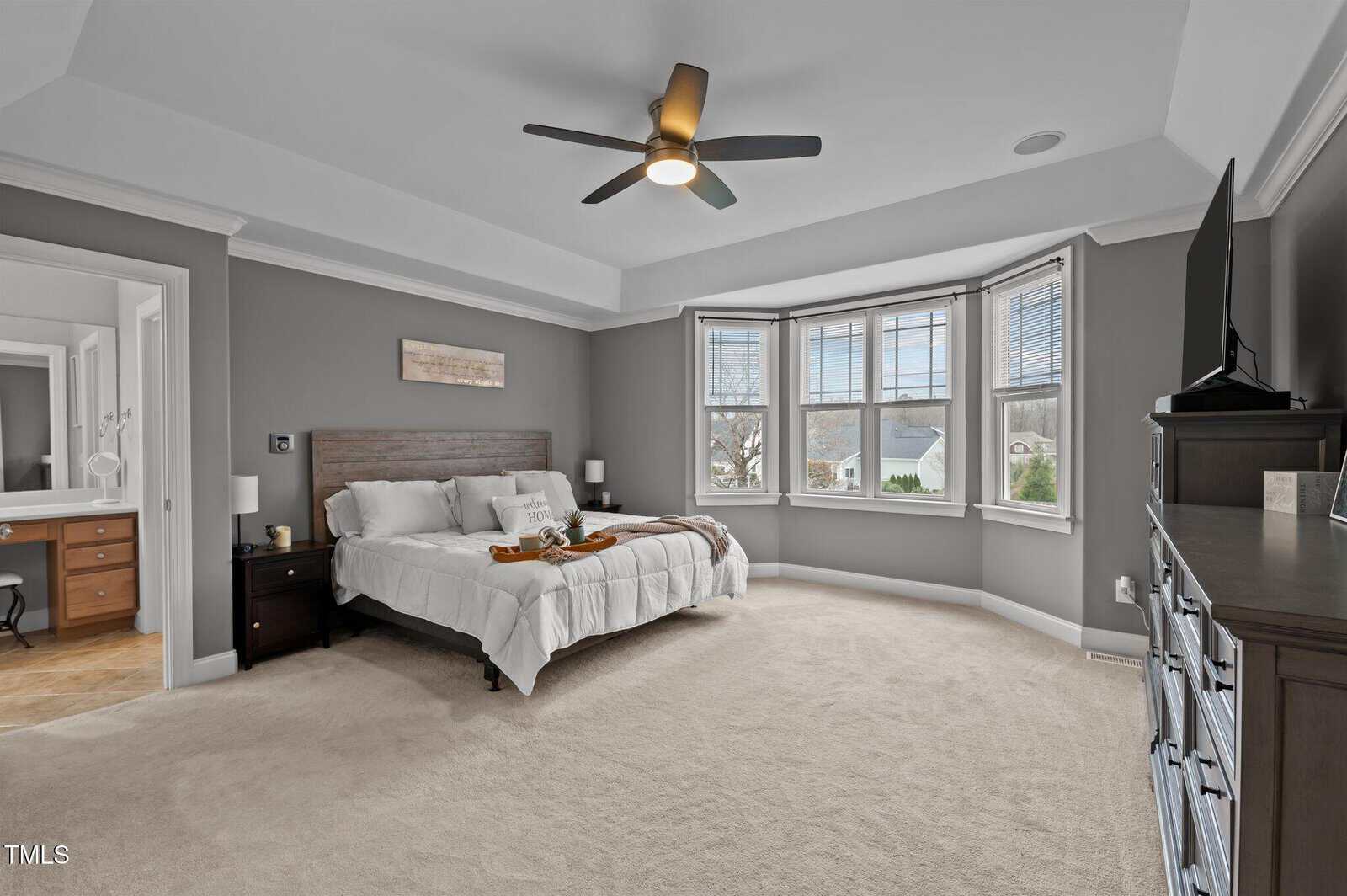 Spacious bedroom with gray walls, a ceiling fan, large bay windows, a wooden bed, two nightstands, and a dresser. An ensuite bathroom with a vanity is visible on the left.