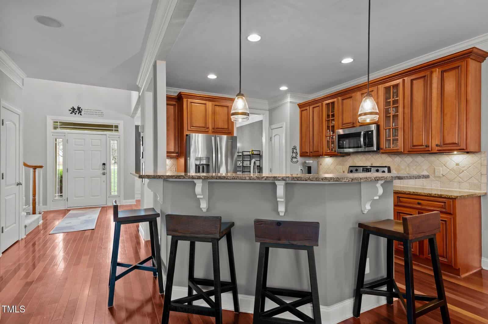 A modern kitchen with wooden cabinets, stainless steel appliances, granite countertops, and three dark wooden barstools at a breakfast bar. Pendant lights hang above the bar and the front door is visible in the background.