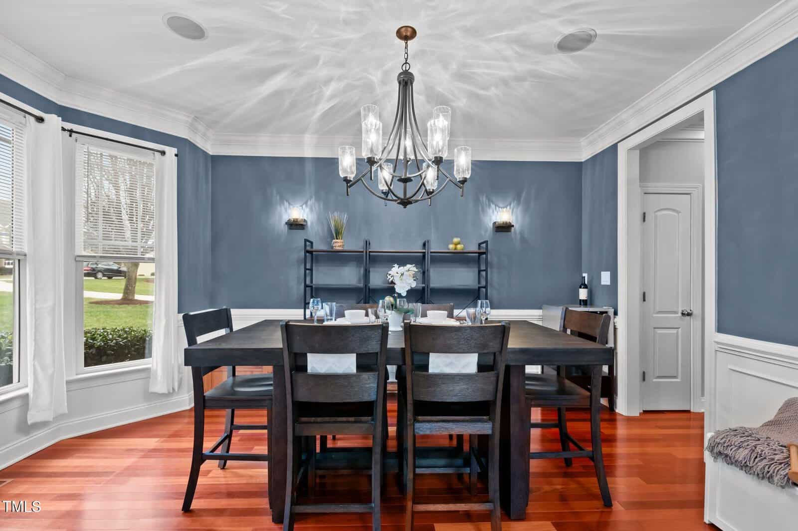 A dining room with blue walls, white trim, and wood floor. A rectangular dark wood table with six chairs is centered under a chandelier. Shelves and a door are in the background; windows with white curtains are on the left.