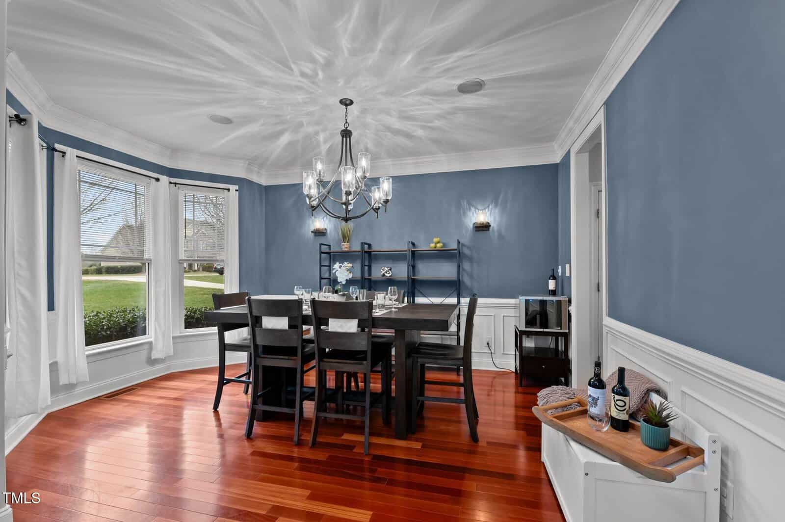 A dining room with blue walls, a large window, hardwood floors, a dark dining table with eight chairs, a chandelier, and a small bench with wine bottles and a plant in the corner.
