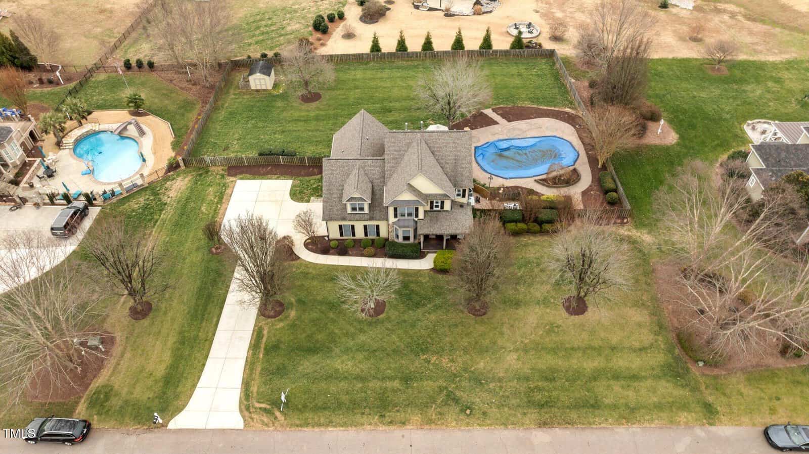 Aerial view of a two-story house with a gray roof, large driveway, landscaped front yard, fenced backyard, and an in-ground pool covered with a blue tarp. Surrounding areas have open grassy spaces and neighboring homes.