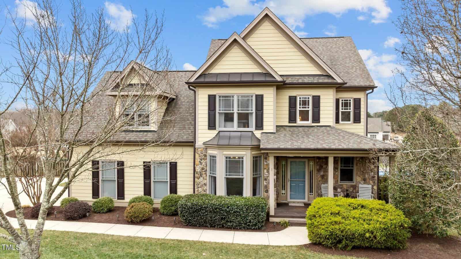 Two-story beige house with brown trim and stone accents, featuring a covered front porch, multiple windows, tidy shrubs, and a well-maintained yard under a partly cloudy sky.