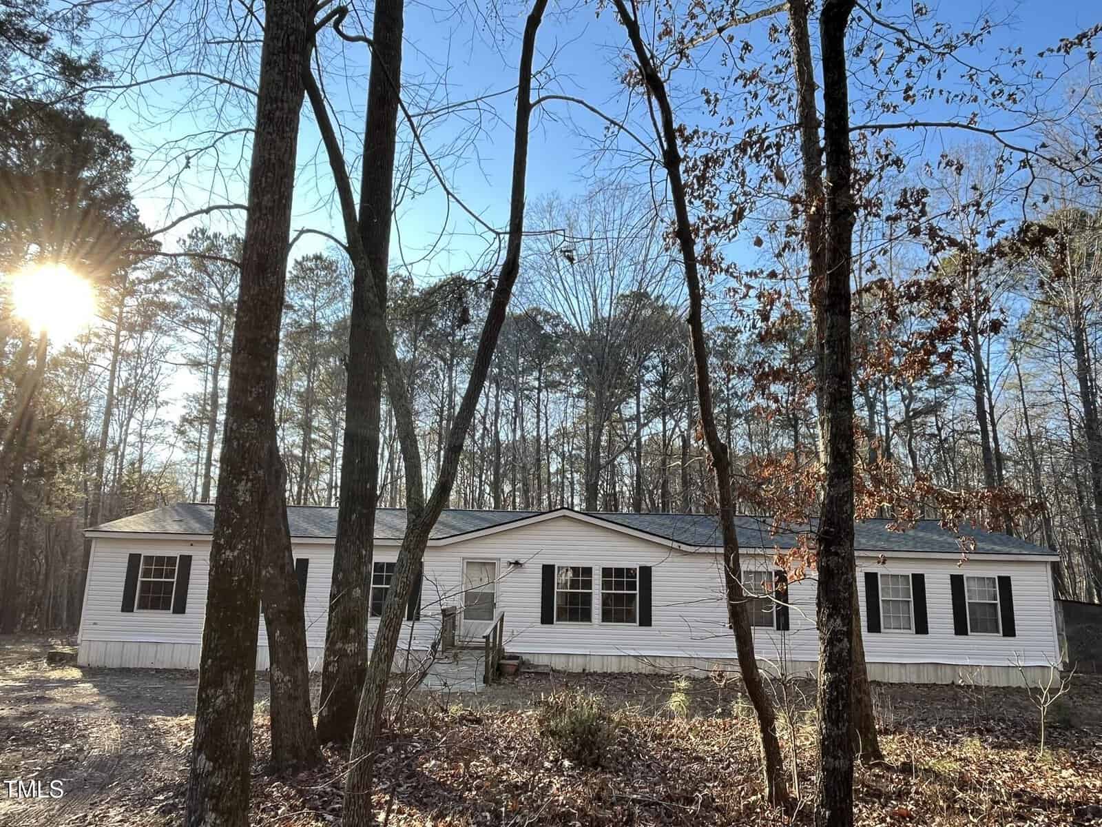 A white manufactured home with black shutters sits in a wooded area. Tall leafless trees surround the home, and sunlight filters through the branches. The ground is covered with dry leaves and patches of dirt.