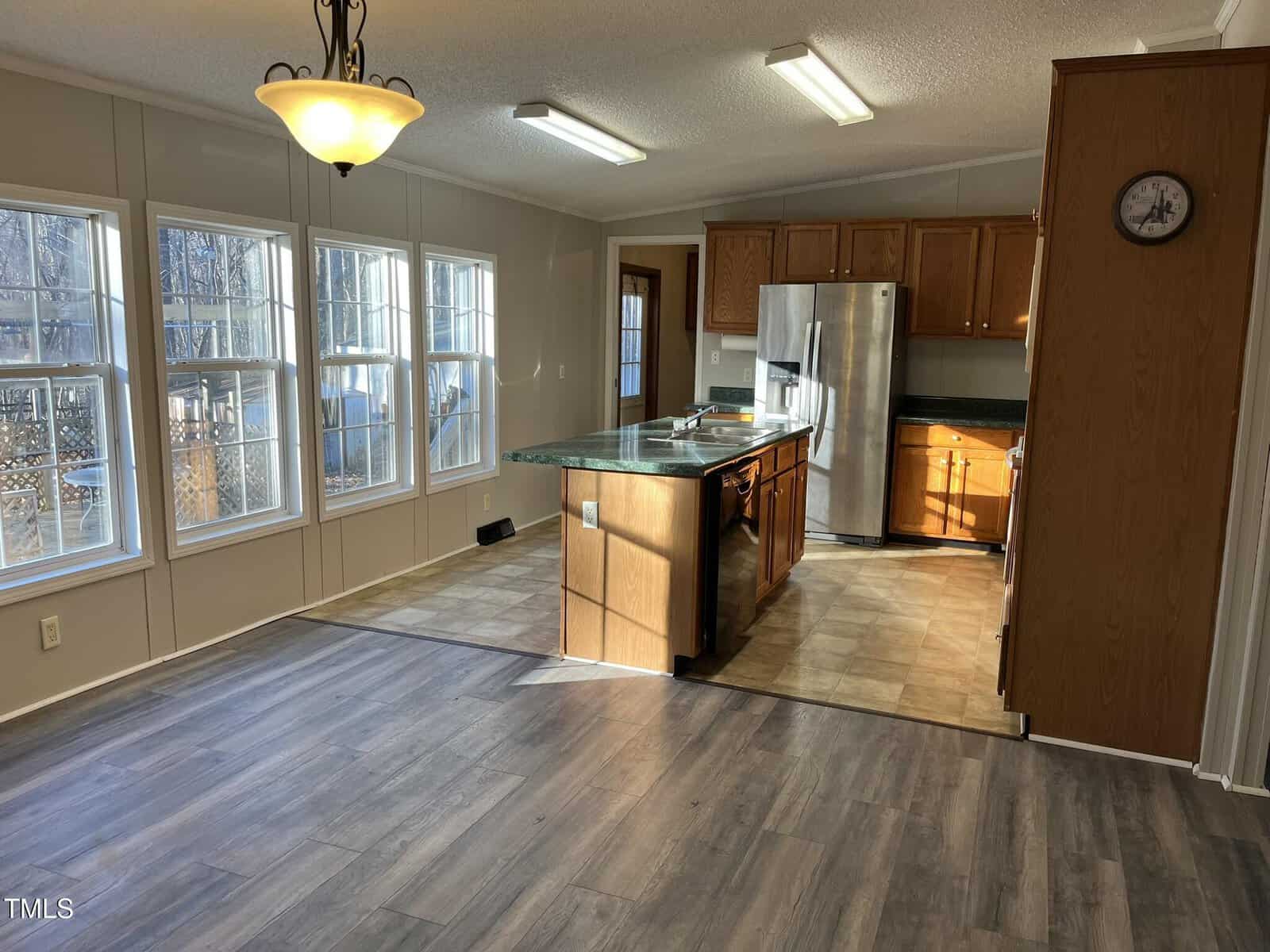 A kitchen with wood cabinets, stainless steel appliances, and a center island opens to a dining area with large windows and wood-look flooring. Overhead lights and natural light brighten the space.