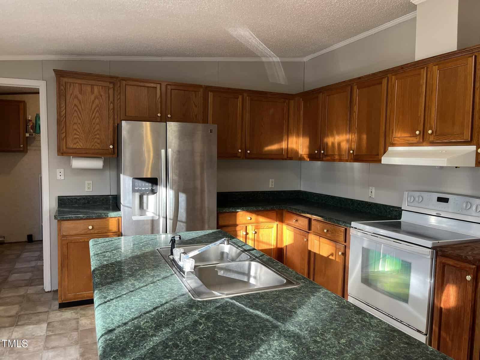 A kitchen with wooden cabinets, dark green marble countertops, a stainless steel refrigerator, a white electric stove with a range hood, and a sink in a central island. Sunlight streams in through an unseen window.