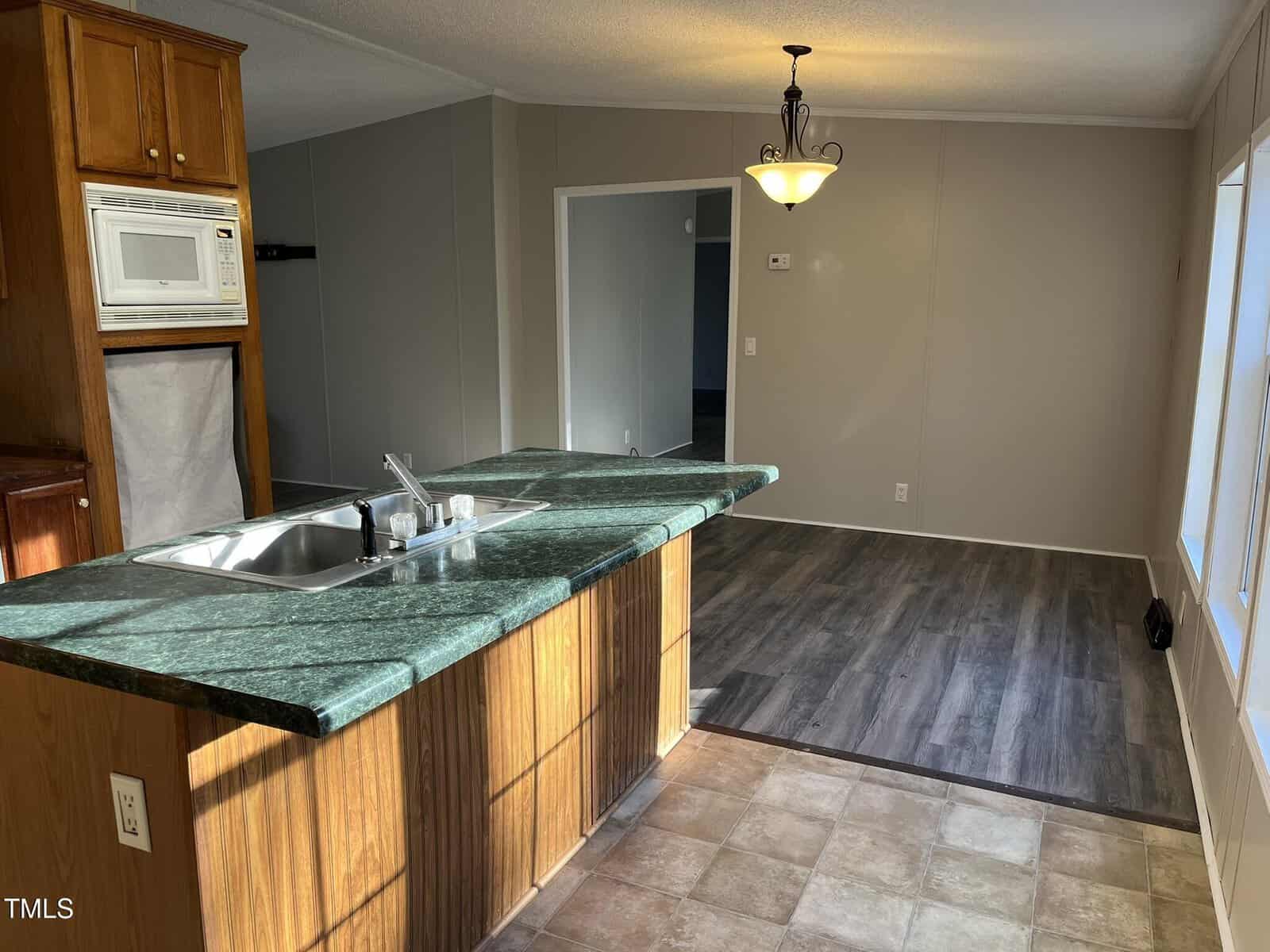 A kitchen with a green marble countertop island, stainless steel sink, and wood cabinets is next to a dining area with wood-look flooring, beige walls, and a ceiling light fixture. Sunlight streams in through a large window.