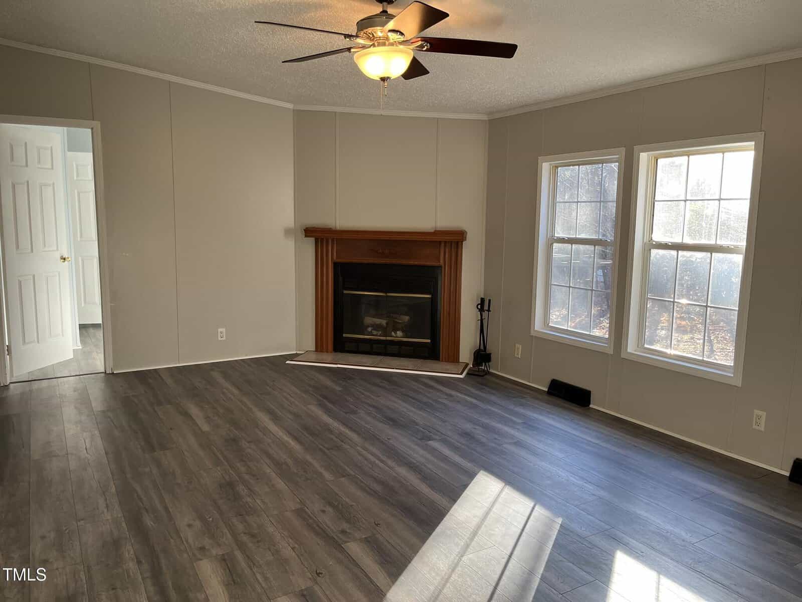 Empty living room with gray wood floors, beige walls, a ceiling fan with light, fireplace with wooden mantel, and three large windows letting in natural light. A doorway leads to another room on the left.