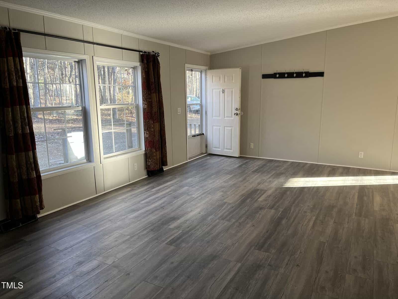 Empty room with wood-style flooring, light gray walls, two large windows with curtains, and a white front door. Sunlight comes in through the windows, highlighting the floor. A simple wall rack is mounted near the door.