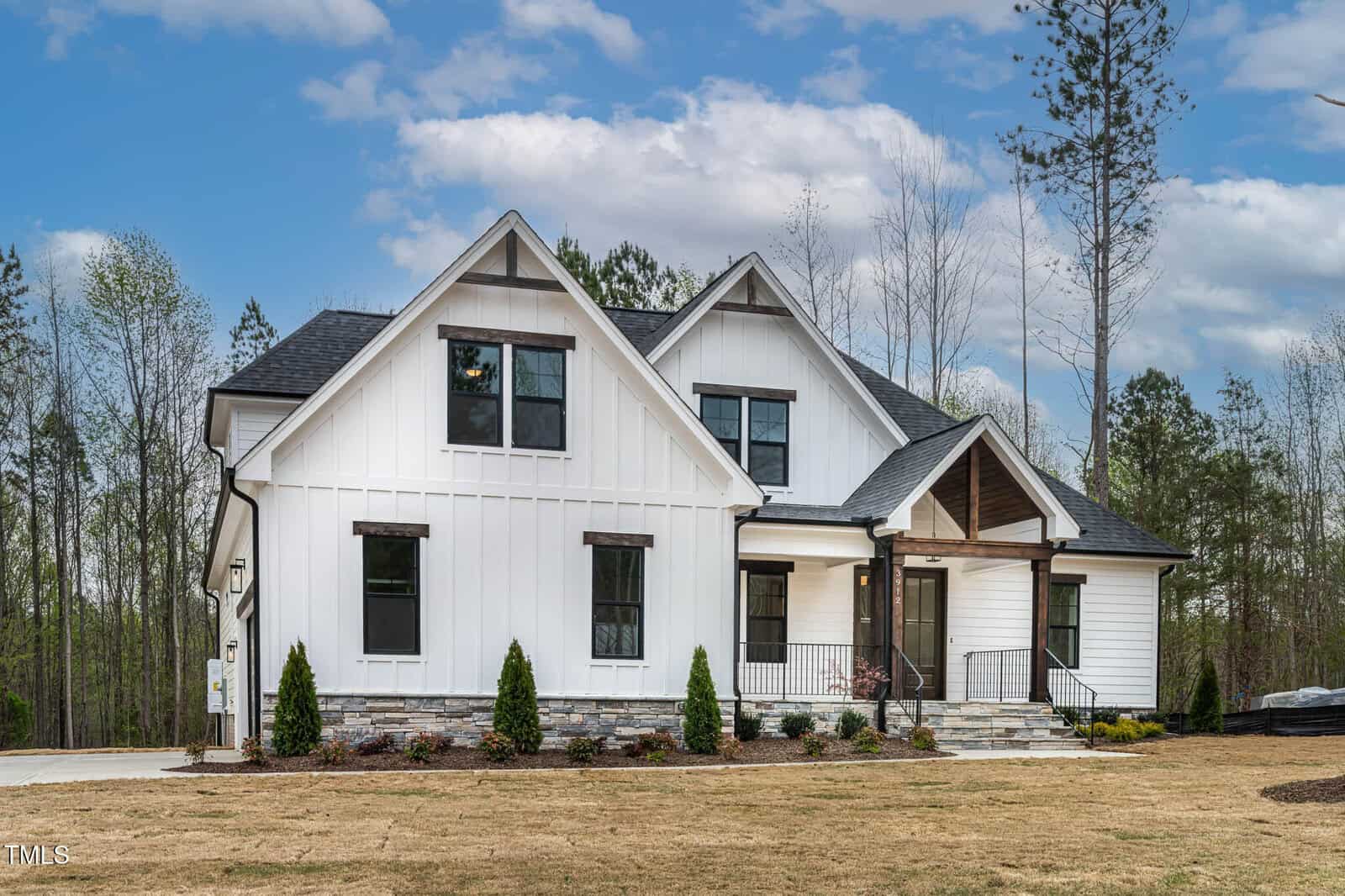 White modern farmhouse with black trim, featuring a gable roof and stone accents. The front has several windows, a porch with stairs, and landscaped shrubs, surrounded by a lawn. Trees are visible in the background under a blue sky with clouds.