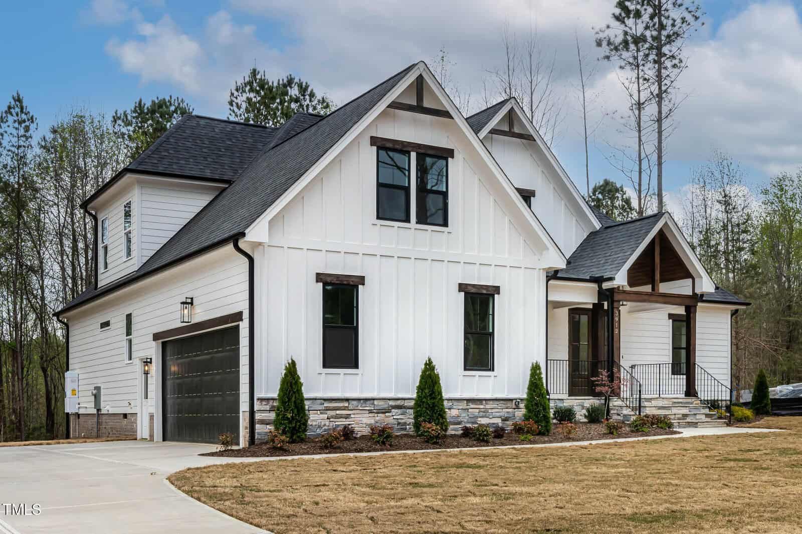 A modern white house with dark gray accents features a gable roof and two stories. The facade has vertical siding and stone trim at the base. There is a two-car garage, a front porch with a gabled entry, and a driveway. The yard is landscaped with shrubs.