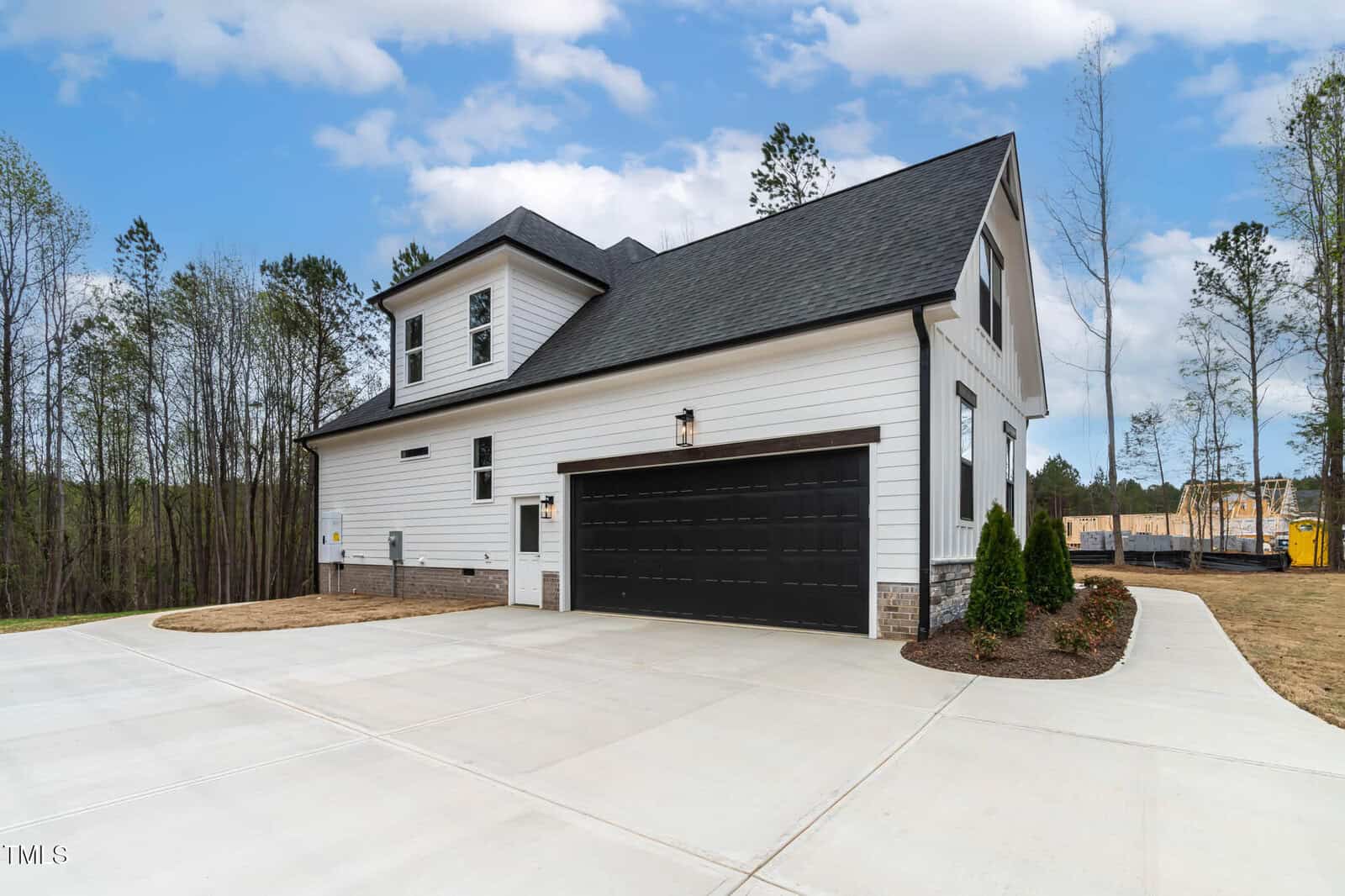 A modern two-story house with white siding and black roof and garage door. It has a paved driveway and a small landscaped area with shrubs. Trees are visible in the background under a partly cloudy sky.