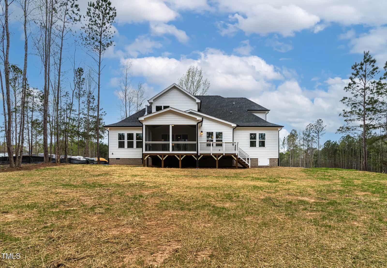 A two-story white house with a dark roof is situated in an open area surrounded by tall, sparse trees. The house features a back porch with stairs leading to a grassy backyard. The sky is partially cloudy.