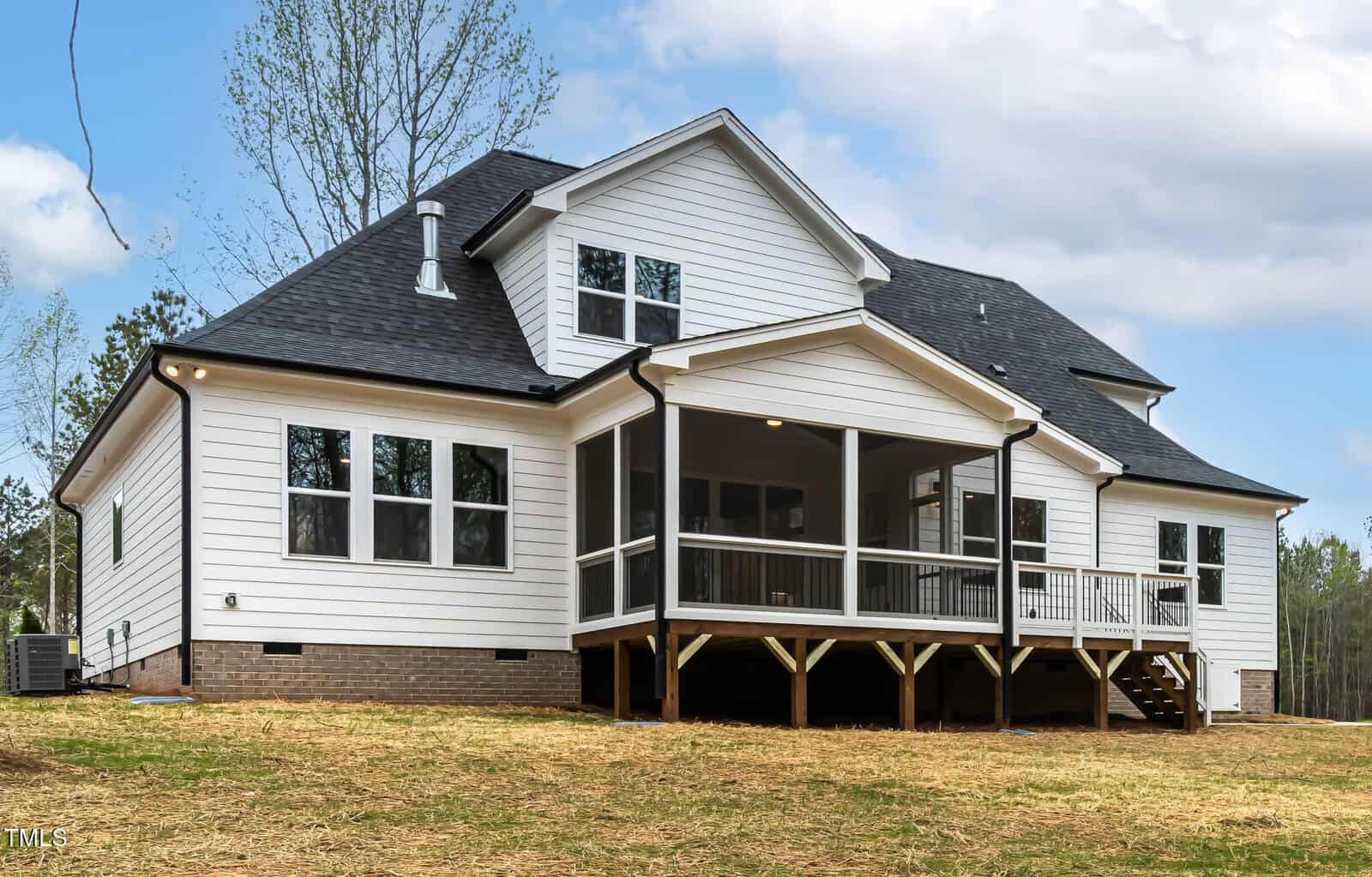 A two-story house with white siding and a black roof is pictured. It features multiple windows and a covered porch with steps leading down to a lawn. The sky is partly cloudy. Trees are visible in the background.