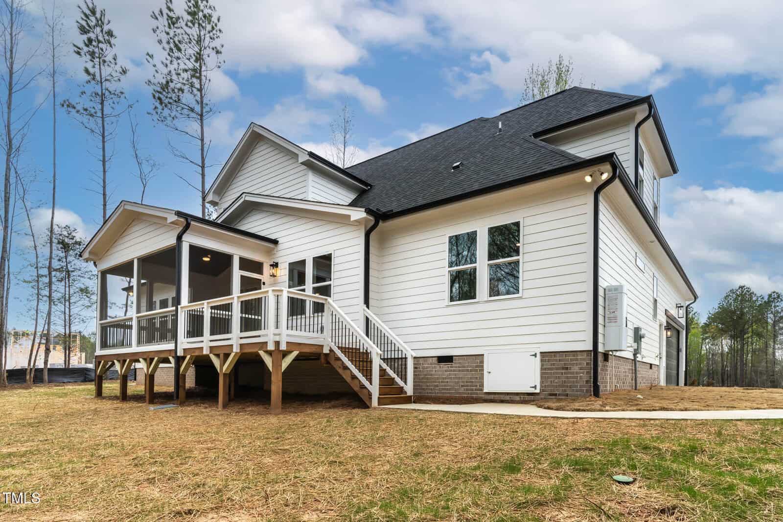 A modern two-story house with white siding and a dark roof. It features a screened porch with stairs leading down to a grassless backyard area. The sky is partly cloudy, and tall, sparse trees are visible in the background.