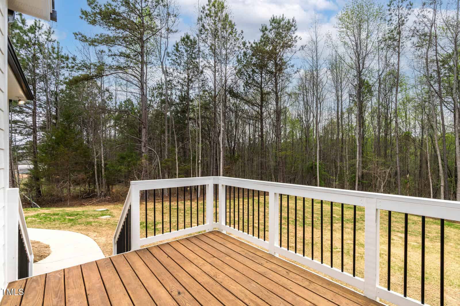 Wooden deck with white railing overlooks a spacious backyard bordered by a line of tall, bare trees. A narrow concrete path leads from the deck to the grassy area. The sky above is partly cloudy.