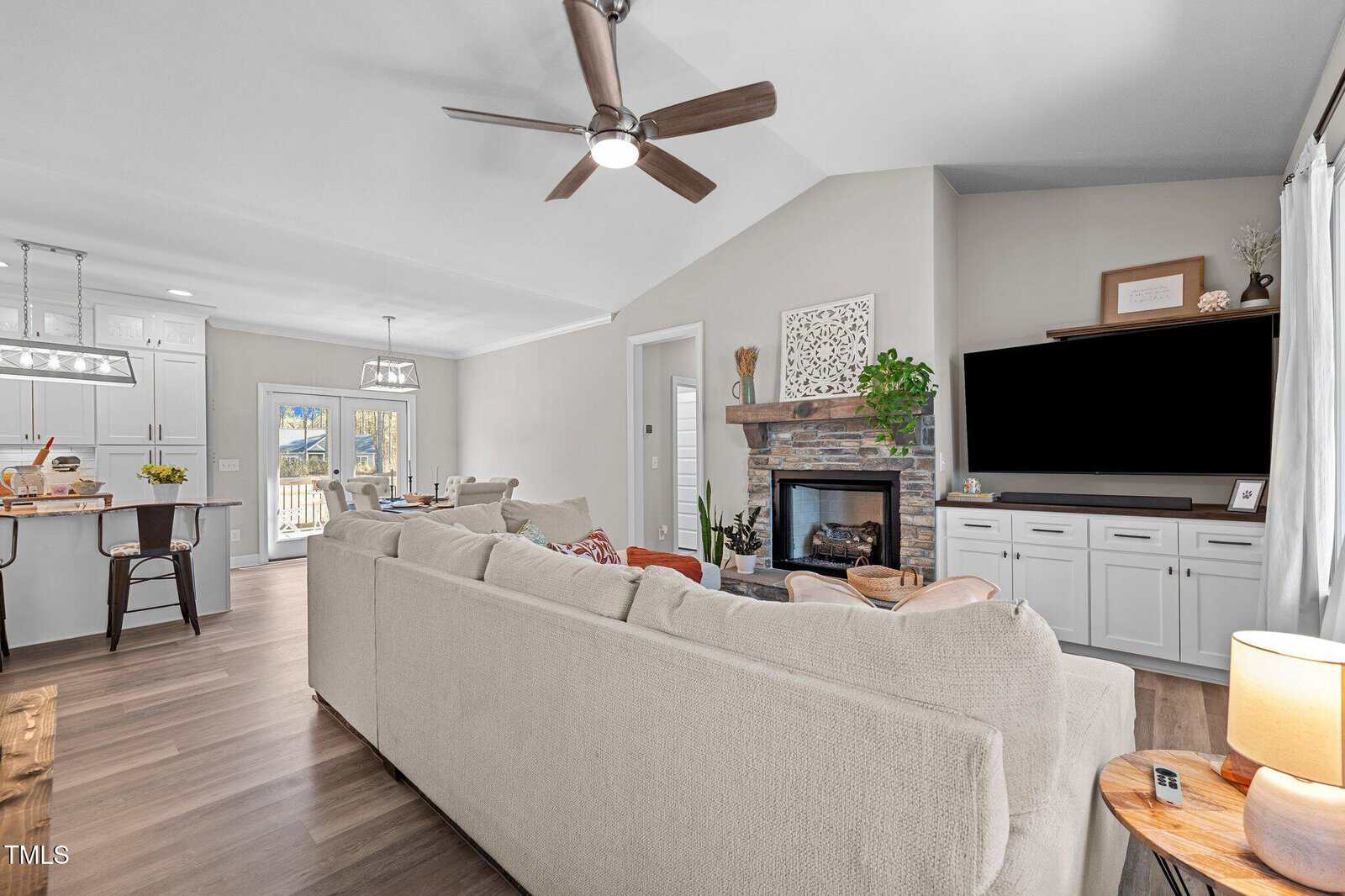 A modern living room with a beige sectional sofa facing a wall-mounted TV above white cabinets. There's a fireplace with a stone surround to the right, and a ceiling fan overhead. The kitchen and dining area are visible in the background.