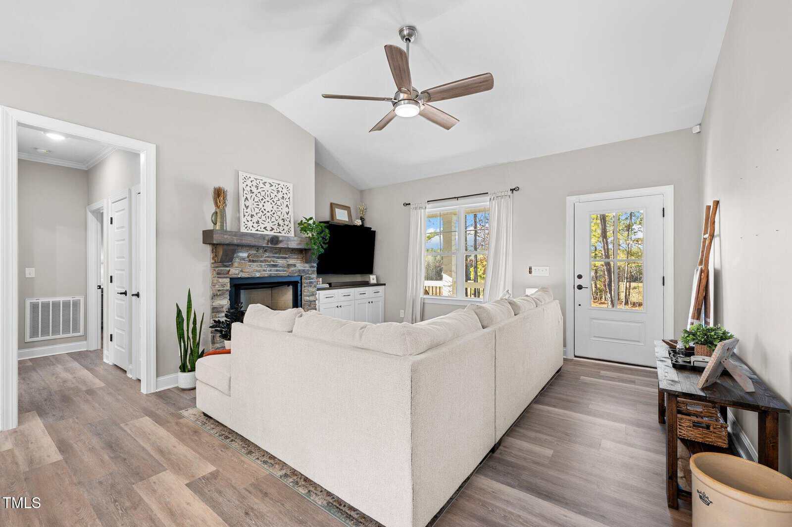 A modern living room with a beige sectional sofa facing a TV on a stone-accented fireplace. There is a ceiling fan above, a door and windows on the right, and potted plants throughout the space. Light wood flooring adds a warm touch.