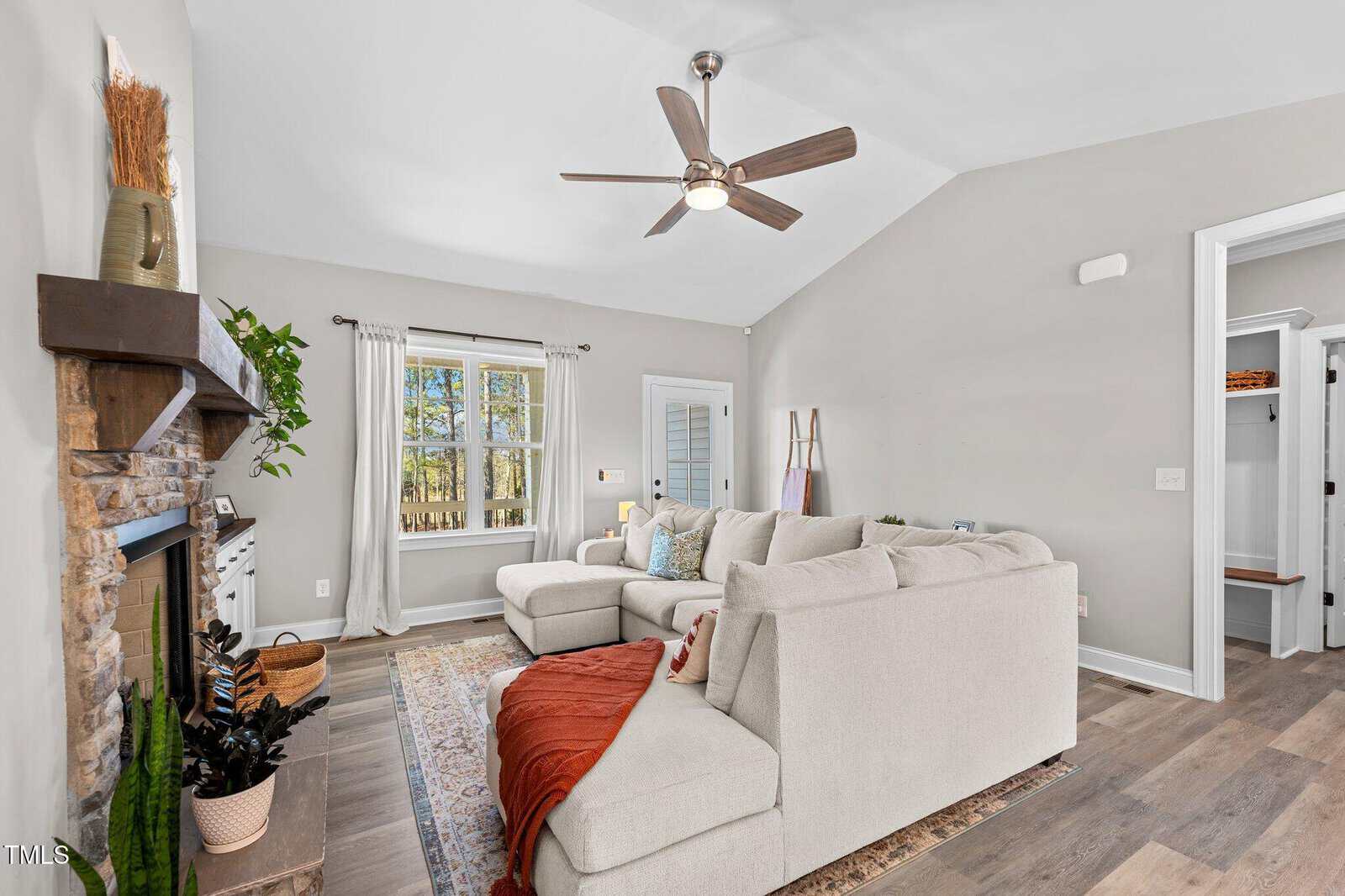 A living room with a beige sectional sofa, wooden coffee table, and a ceiling fan. There's a stone fireplace with a wooden mantel, a large window with curtains, and a light-colored rug on the floor. A doorway leads to another room.