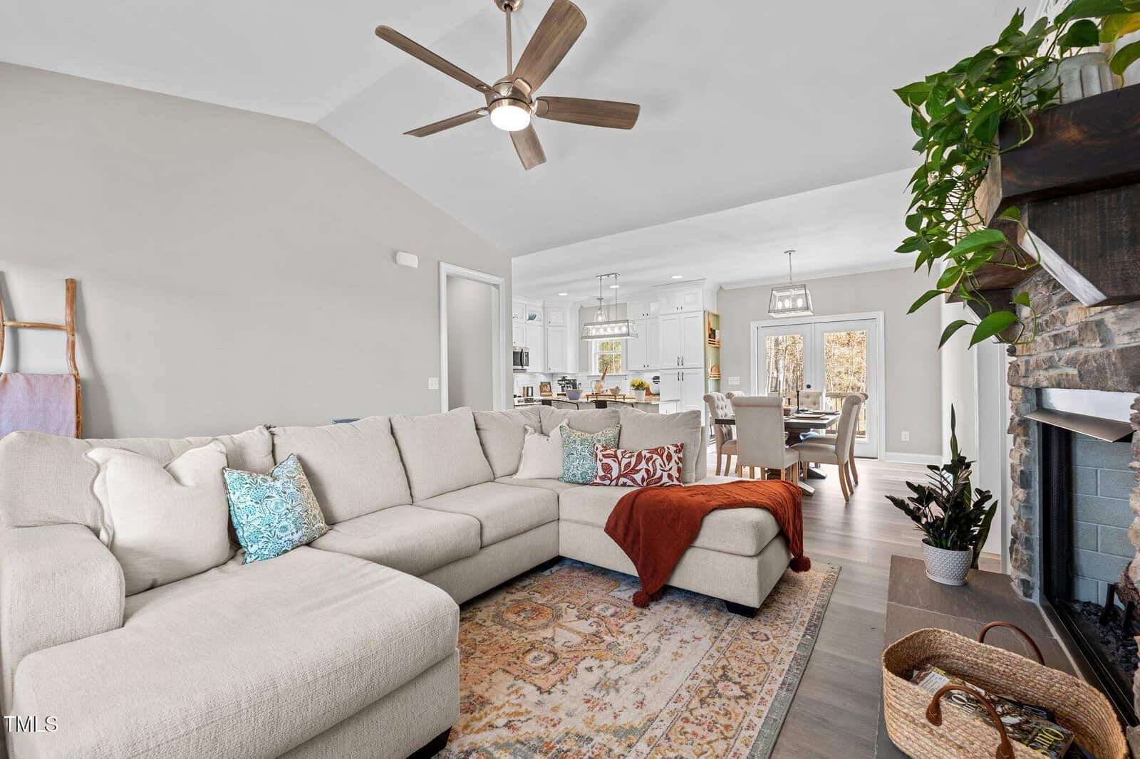 A spacious living room with a beige sectional sofa, colorful cushions, and a red throw blanket. A ceiling fan is above. The room extends into a white open kitchen and dining area. A stone fireplace and a plant are visible on the right.
