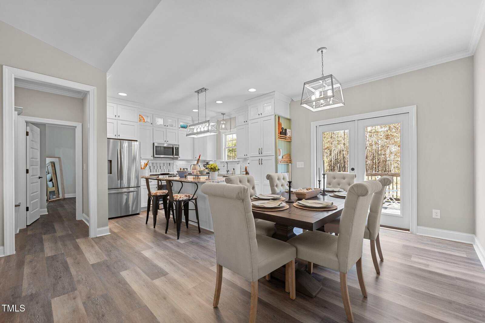 A modern kitchen and dining area with hardwood floors. The kitchen has white cabinets, stainless steel appliances, and a central island with black stools. The dining area features a rectangular table with beige cushioned chairs. A glass door leads outside.