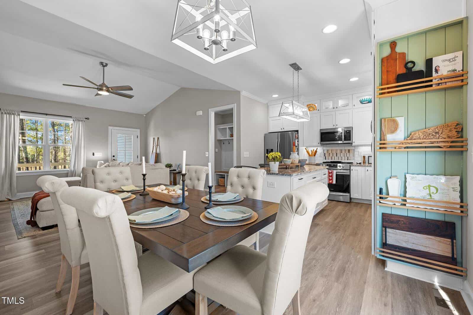 A modern open-concept living room and kitchen area with light-colored walls and wood flooring. The dining table has six upholstered chairs and is set with plates and candles. A ceiling fan, pendant lights, and built-in shelves add to the decor.