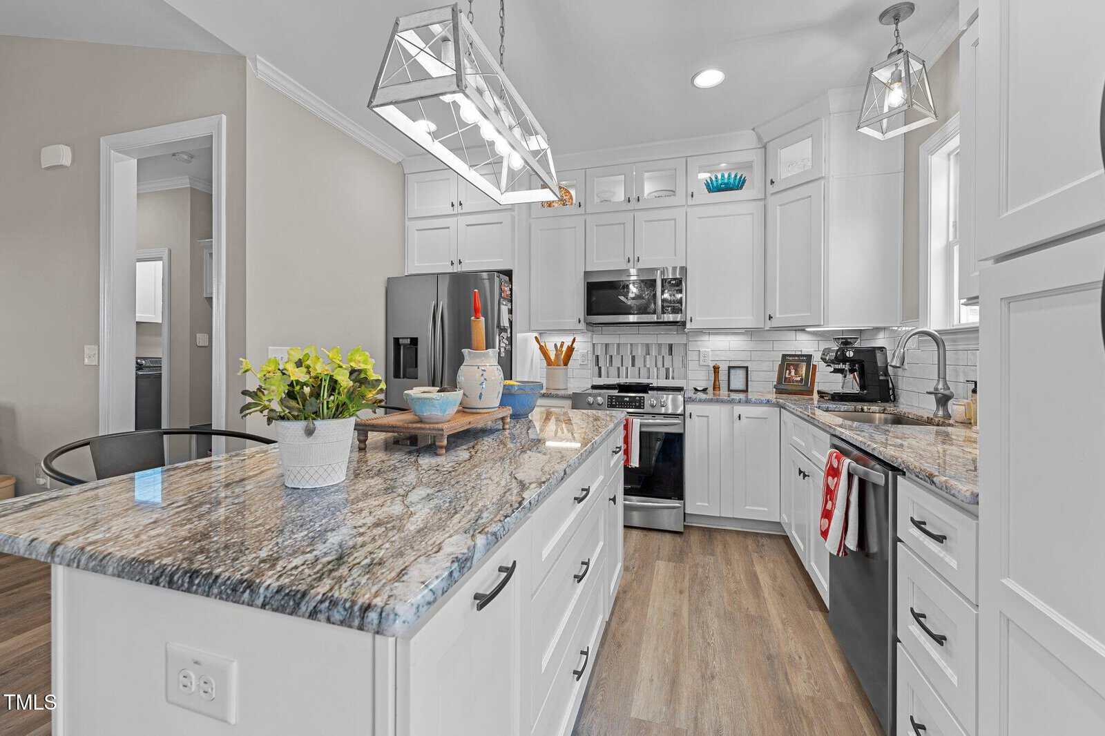A modern kitchen featuring white cabinets, stainless steel appliances, and a marble countertop with a built-in island. The island displays a potted plant and kitchenware. Pendant lights hang above, illuminating the hardwood floor.
