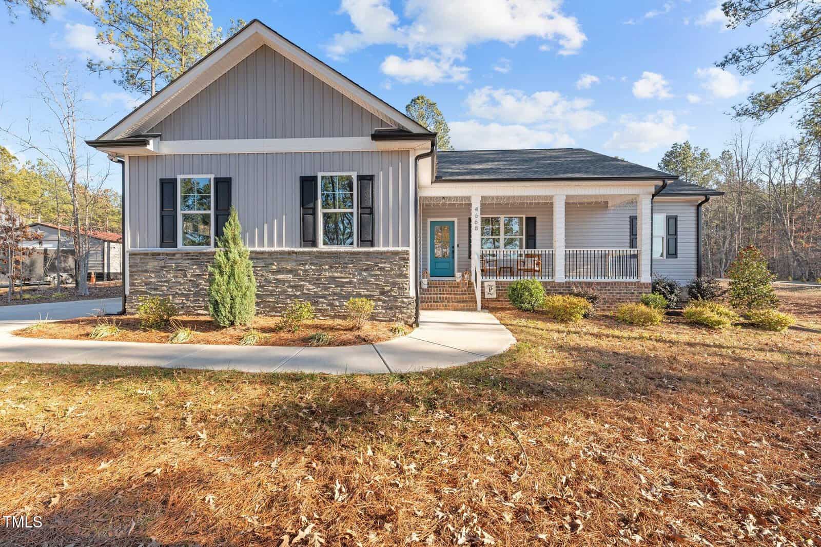 Single-story house with gray siding and stone accents, featuring a front porch with a teal door, set on a grassy yard with trees in the background. The sky is clear with a few clouds.