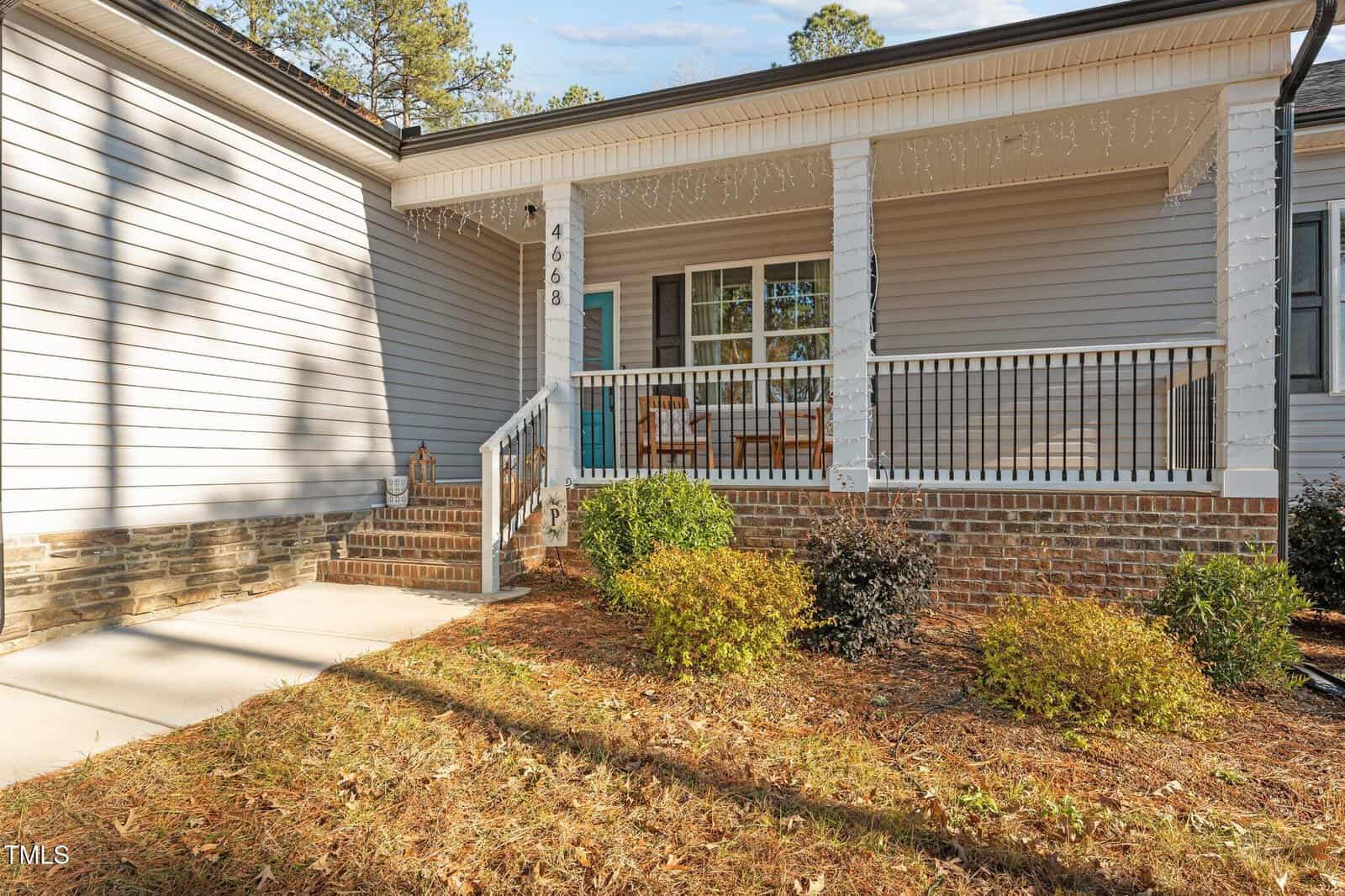 A house with gray siding features a porch adorned with string lights. The porch has a wooden railing and two chairs. The pathway leads to the front door, which is painted blue. Shrubs and pine straw are planted in the front yard.