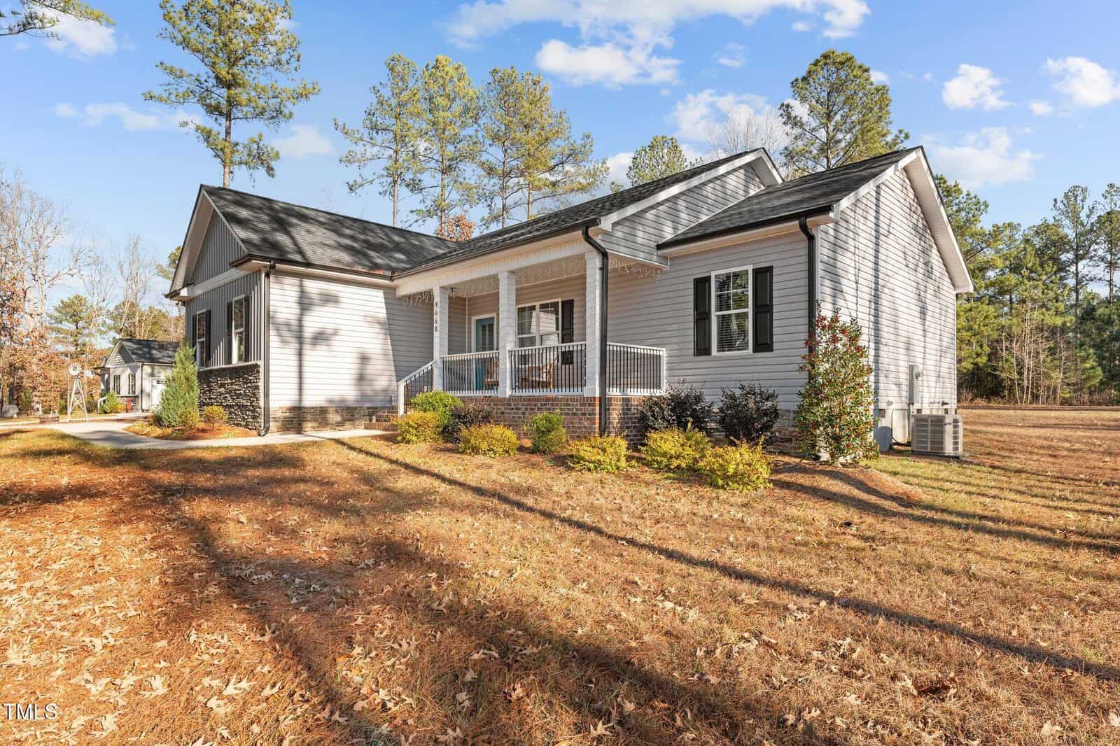 A single-story house with light gray siding and a dark roof is surrounded by grass and trees. A small porch with white railings is at the entrance. The sky is clear with a few clouds.