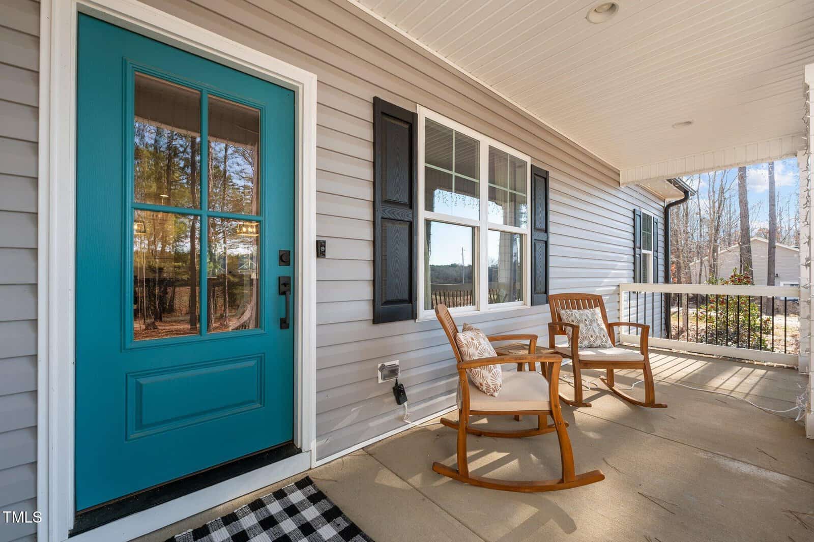 A front porch with two wooden rocking chairs and a teal-colored door. The house has gray siding and black shutters. A checkered doormat lies near the door. Trees and a neighboring house are visible in the background.