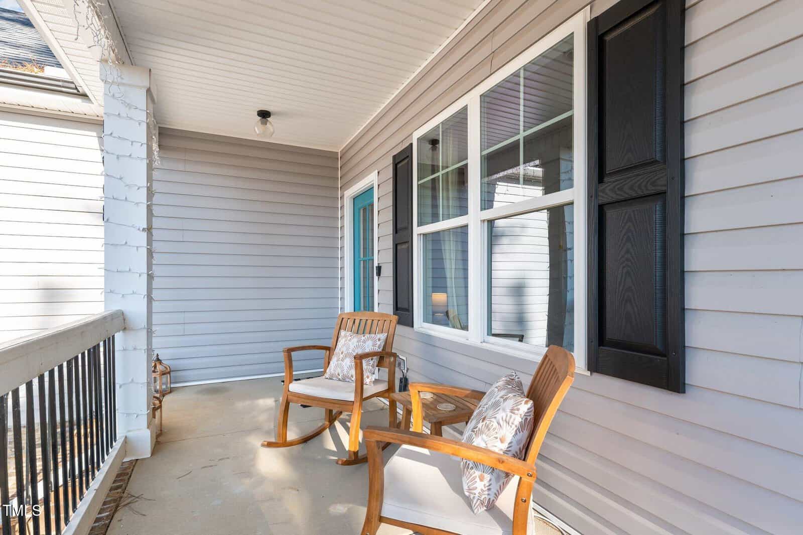 A porch with two wooden chairs and cushions on a concrete floor. The exterior wall is gray with white-trimmed windows and black shutters. The ceiling has a single light fixture, and a blue door is partially visible.