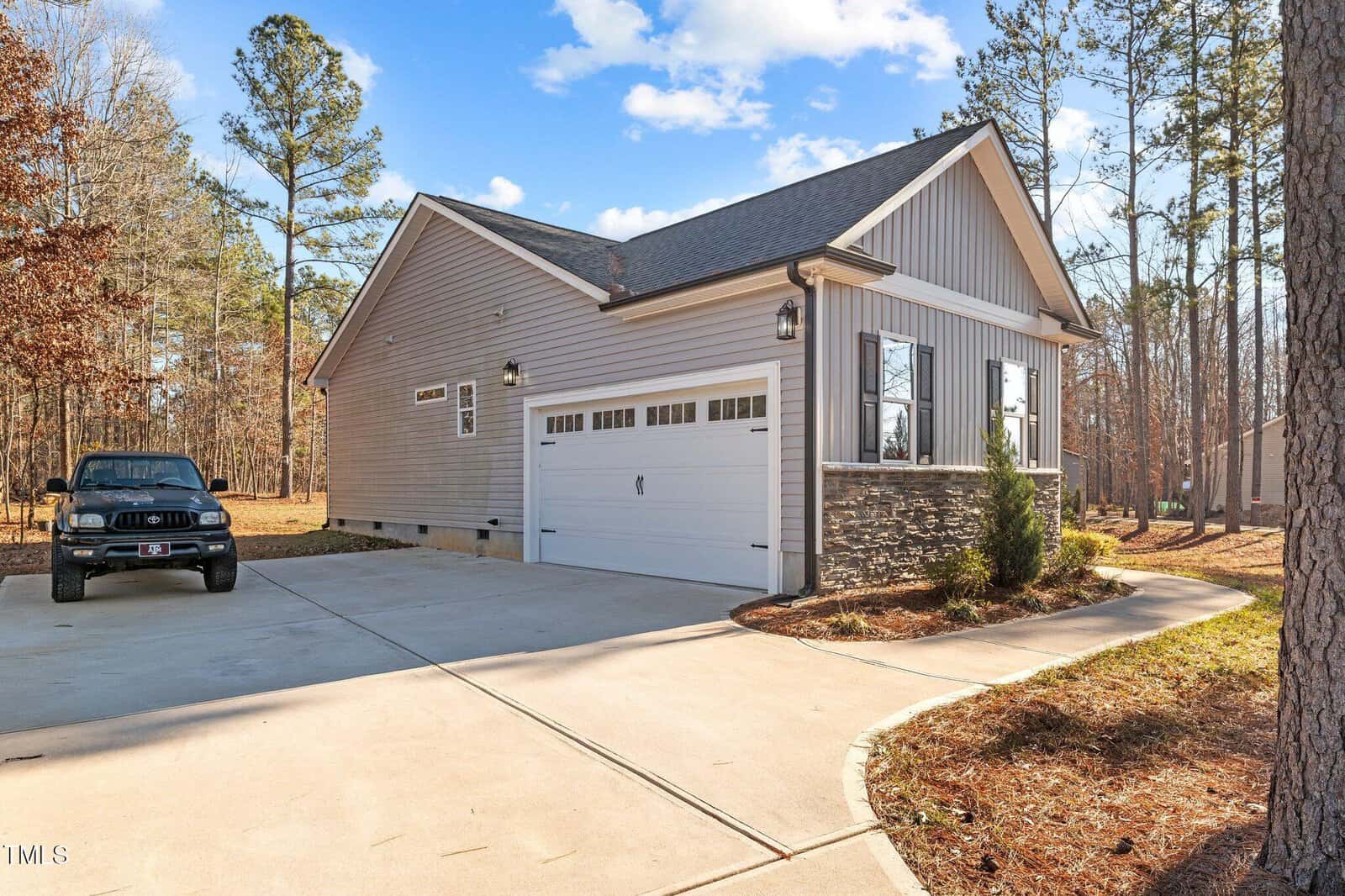 Single-story house with gray siding and stone accents, featuring a two-car garage. The driveway leads to the garage, with a black SUV parked on the left. The house is surrounded by trees and has a clear blue sky above.