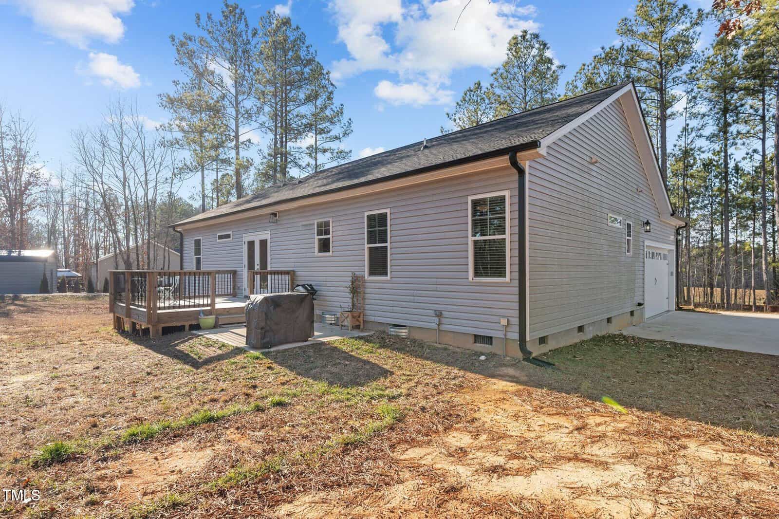 A gray single-story house with a wooden deck and a hot tub on the side. The house is surrounded by trees and a grassy yard, with a clear blue sky above. The building has multiple windows and a two-car garage.