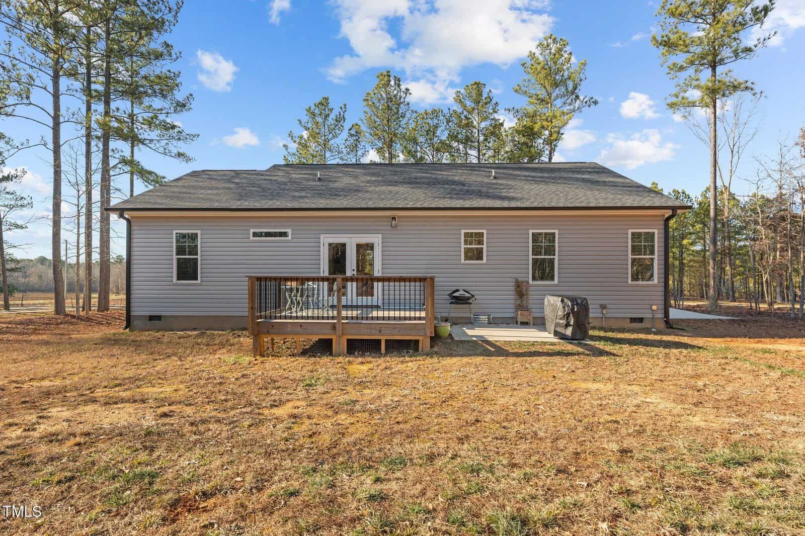 A single-story house with gray siding and a dark roof. The backyard features a small wooden deck with a grill and patio furniture. Surrounding the house are tall pine trees under a partly cloudy sky. The ground is covered with dry grass.