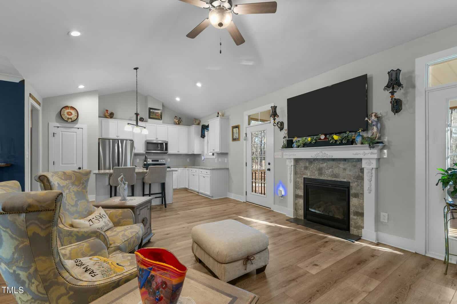 Bright living room with wood floors, a fireplace, wall-mounted TV, armchair, ottoman, open kitchen area, bar stools, stainless steel appliances, and skylight ceiling fan. Decor items add a cozy, welcoming feel.