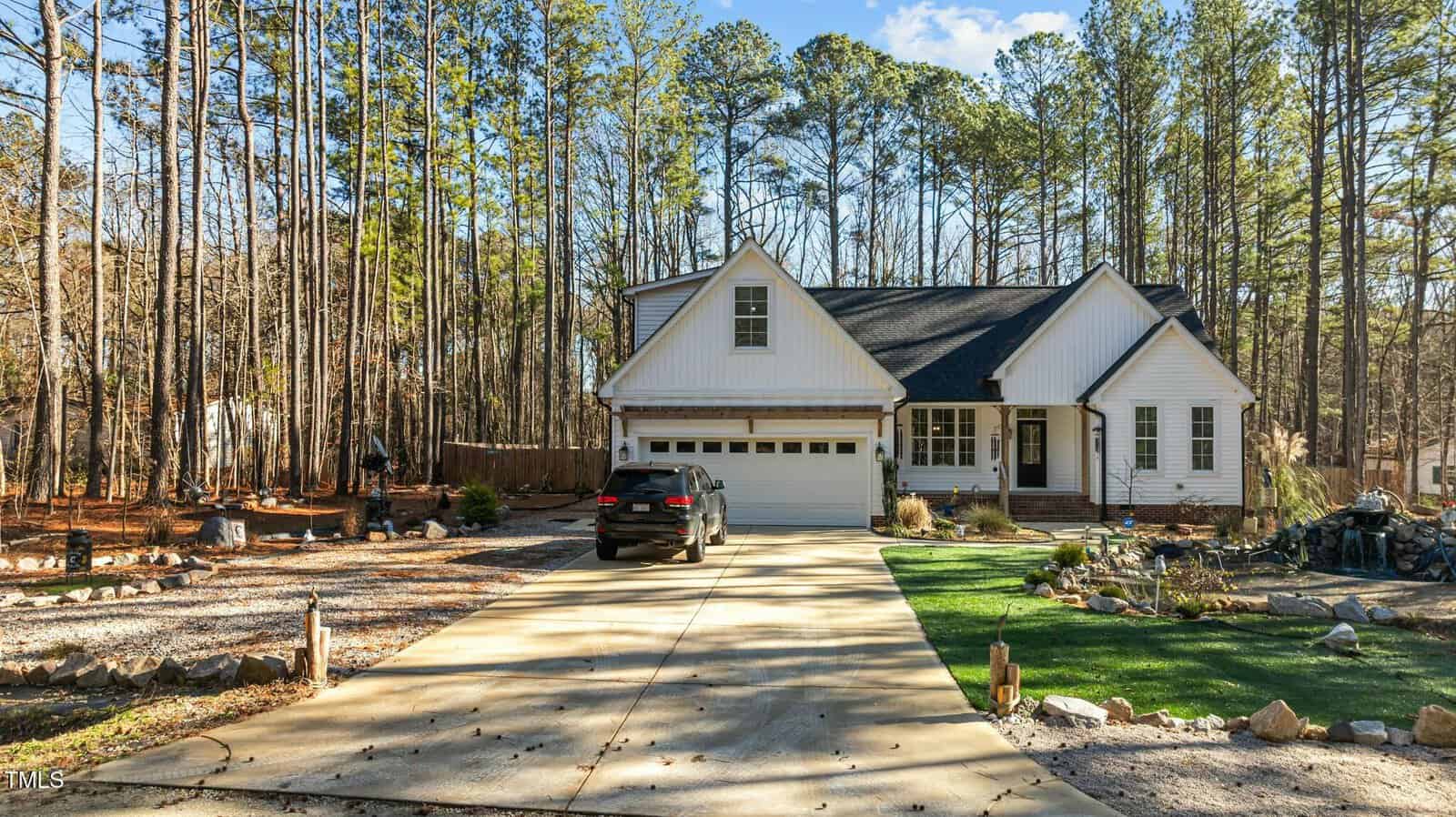 A black SUV is parked in the driveway of a white, single-story house with a front porch and attached garage. The yard has patches of grass, landscaping rocks, and is surrounded by tall pine trees.