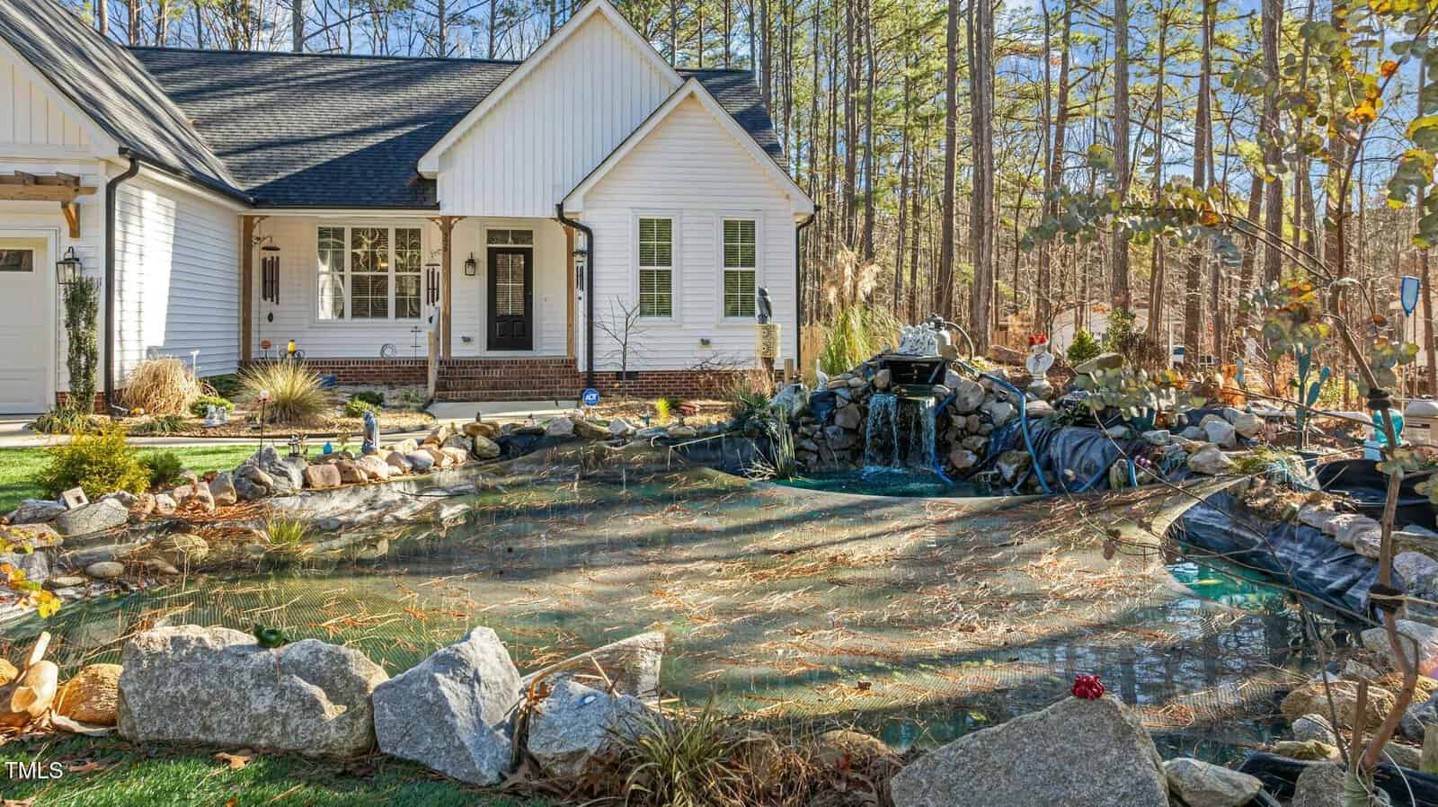 A white house with black trim sits next to a garden pond surrounded by rocks, plants, and pine trees. Water flows over a rock waterfall feature into the pond, which has some leaves and debris on the surface.