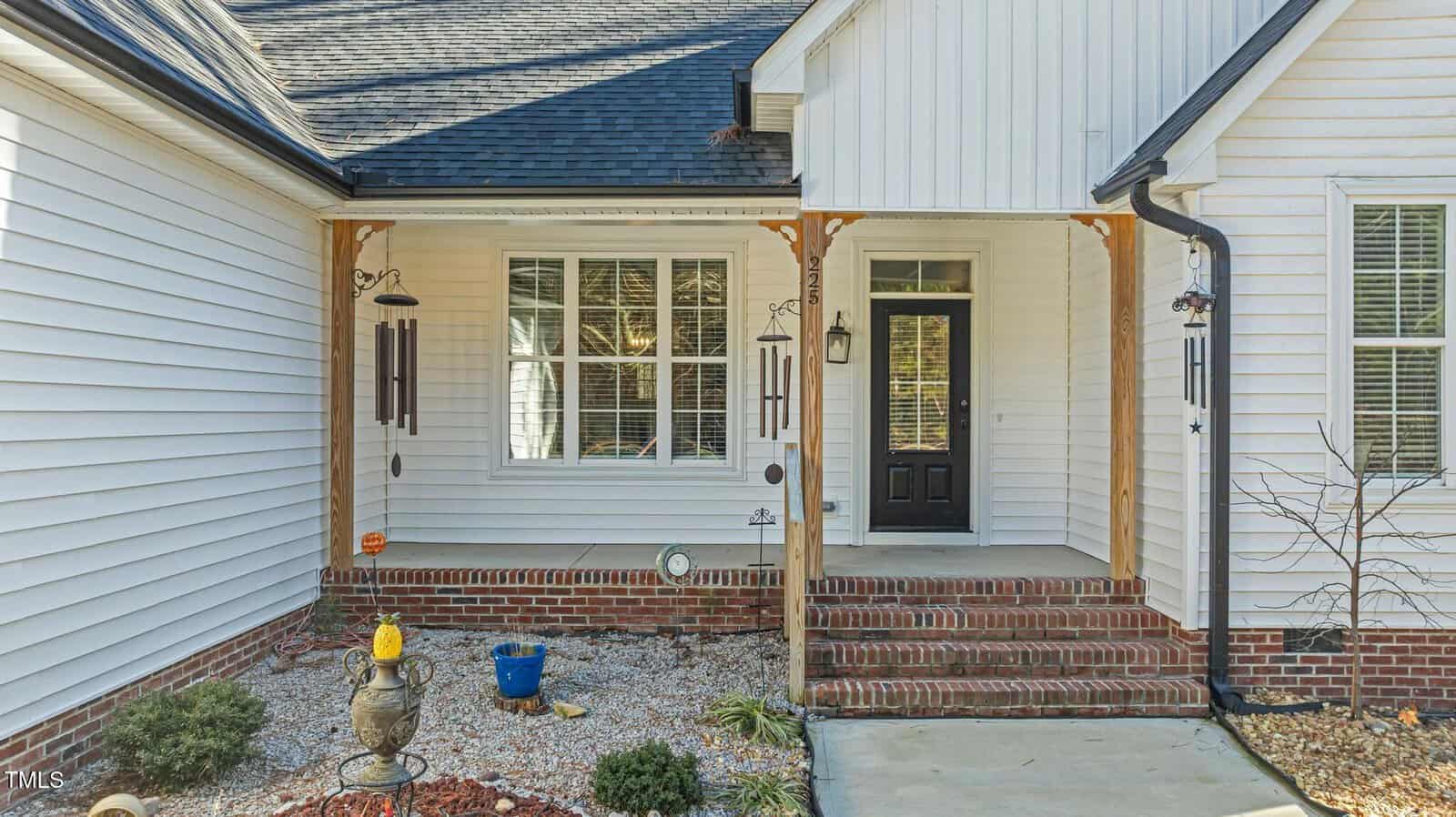 Front porch of a white house with brick steps, wooden posts, wind chimes, potted plants, and a black front door. Landscaping includes rocks, mulch, and a ceramic birdbath in the foreground.