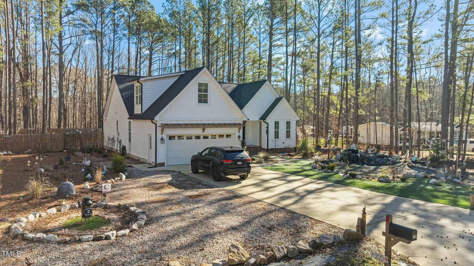 A white house with a black-roofed garage sits among tall trees. A black car is parked in the driveway, and the yard features garden beds with rocks and a small pond. Sunlight casts long shadows across the scene.