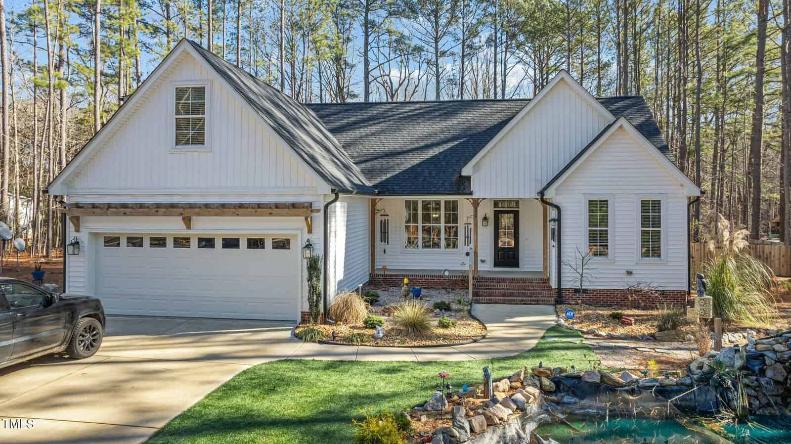 A white, single-story house with a two-car garage, black roof, and brick accents. The front yard features a landscaped garden, pond, and driveway, surrounded by tall trees.